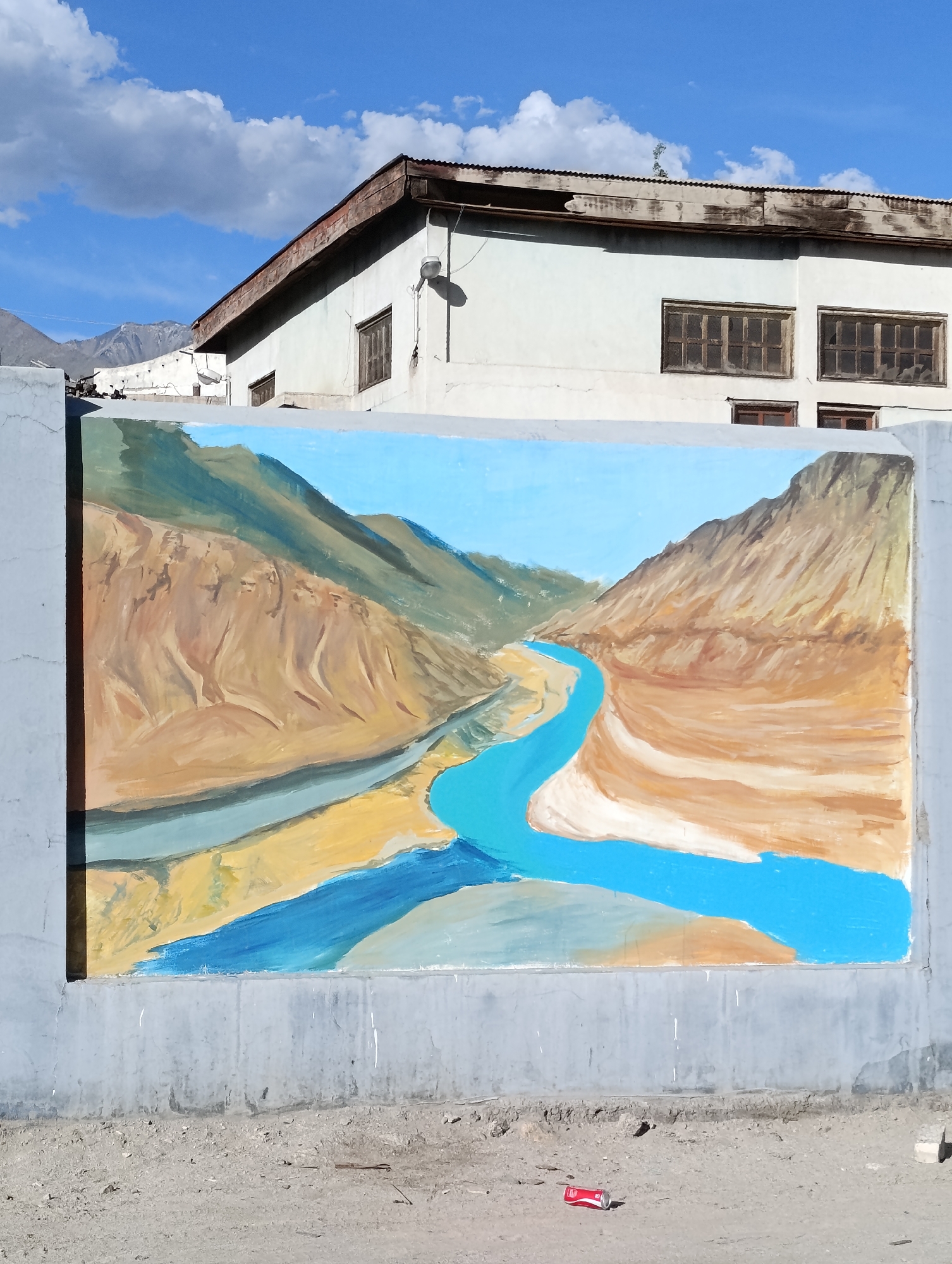 This image depicts a mural painted on a concrete wall in Leh, India. The mural shows a vibrant landscape with a bright blue river winding through arid, mountainous terrain. The mountains are rendered in shades of brown and green, with a clear blue sky overhead dotted with white clouds.
Behind the mural, a light-colored building with a corrugated metal roof is visible. The building has several windows with dark wooden frames. The wall itself is weathered, with some areas showing signs of wear and tear. The ground in front of the wall is a mix of dirt and gravel, with a discarded red and white soda can lying on the ground. The scene suggests an outdoor environment in a dry, possibly high-altitude region.
