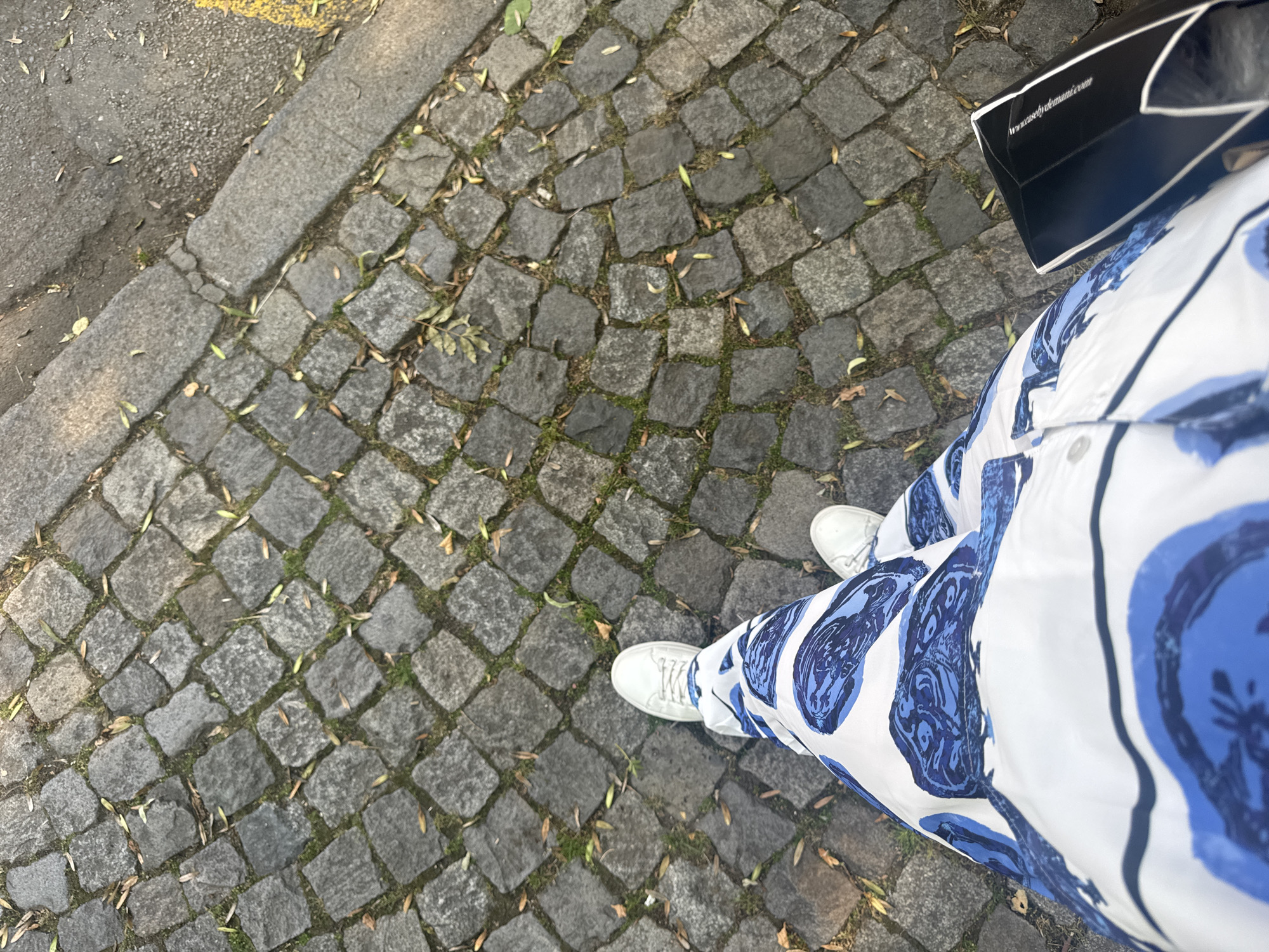 A person is walking on a cobblestone street in Sofia, Bulgaria, as indicated by the architectural style of the paving stones and the general urban environment. The person is wearing white sneakers and a long white dress with a blue abstract pattern. They are carrying a black box bag. The street is lined with a curb separating it from a paved area. Fallen leaves are scattered on the cobblestones, suggesting an autumn season. The lighting indicates it is daytime, with sunlight casting shadows on the ground.