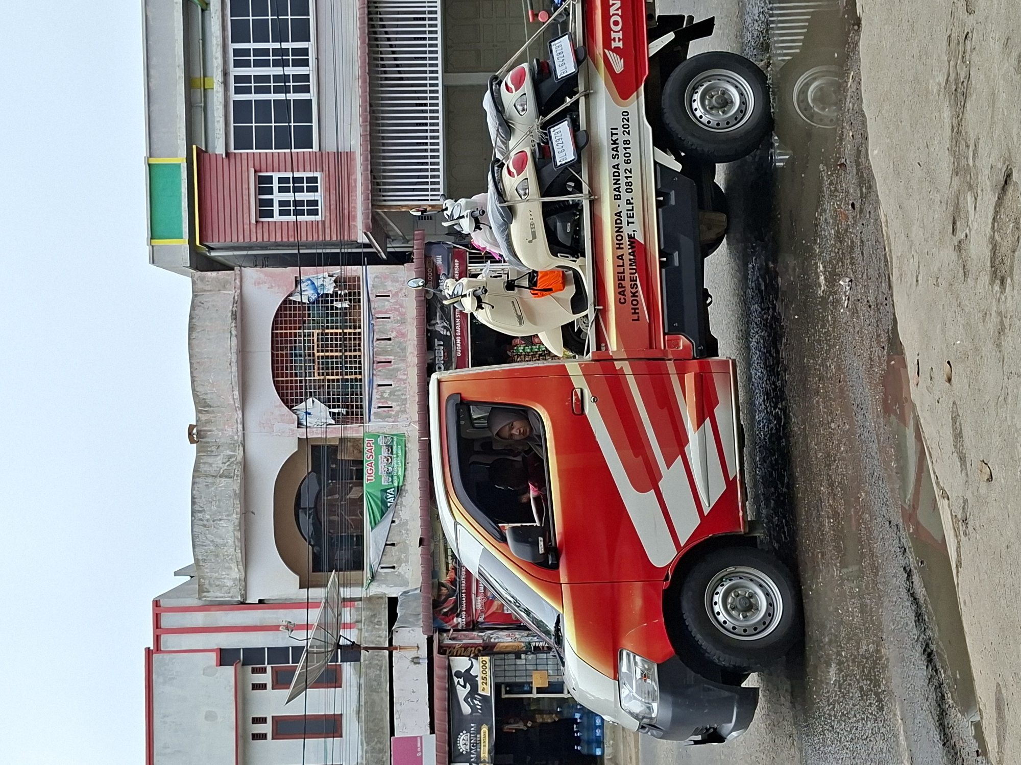 A red and white pickup truck is parked on a wet street in Lhokseumawe, Indonesia. The truck is loaded with two light-colored scooters in the back. The side of the truck has writing that reads "CAPELLA HONDA - BANDA SAKTI LHOKSEUMAWE, TELP. 0812 6018 2020" and a Honda logo. A woman wearing a headscarf is in the driver's seat. In the background, there are several multi-story buildings with storefronts. The sky is overcast.