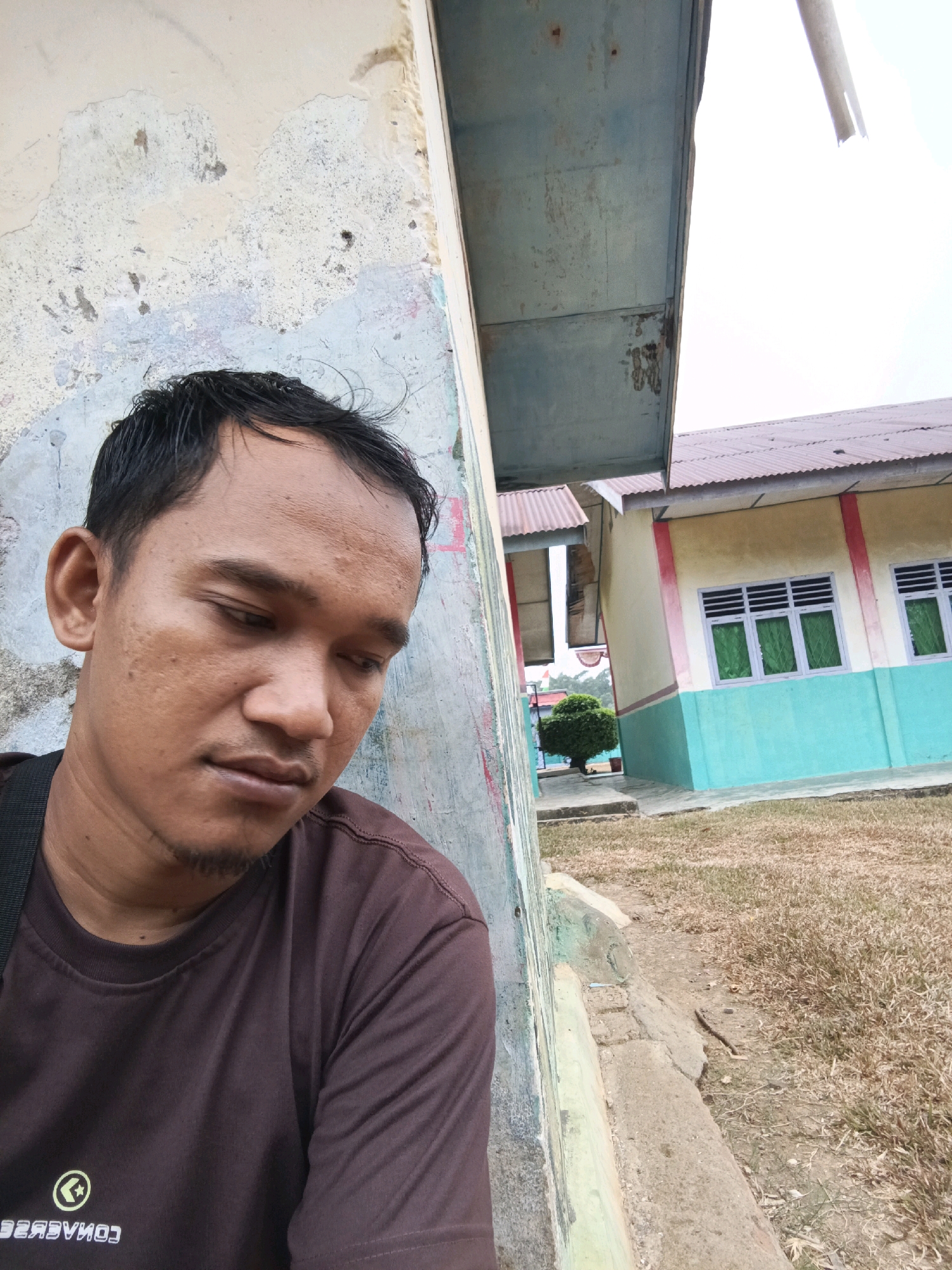 A medium shot selfie shows a man with short, dark hair and light brown skin leaning against a weathered, light-colored wall. He is wearing a dark brown t-shirt with a white logo on the left chest. His head is slightly tilted down and to the right, with his gaze directed downwards. He has a light stubble on his chin and upper lip. Behind him, to the right, is a building with a yellow and turquoise facade, featuring windows with green blinds. The building has a corrugated metal roof. The ground in front of the building is covered with dry grass and a concrete pathway. The overall environment appears to be outdoors in a somewhat rural or neglected setting, possibly a school or community building in a location like Wonosari, Indonesia, given the architectural style and vegetation. The sky is overcast.