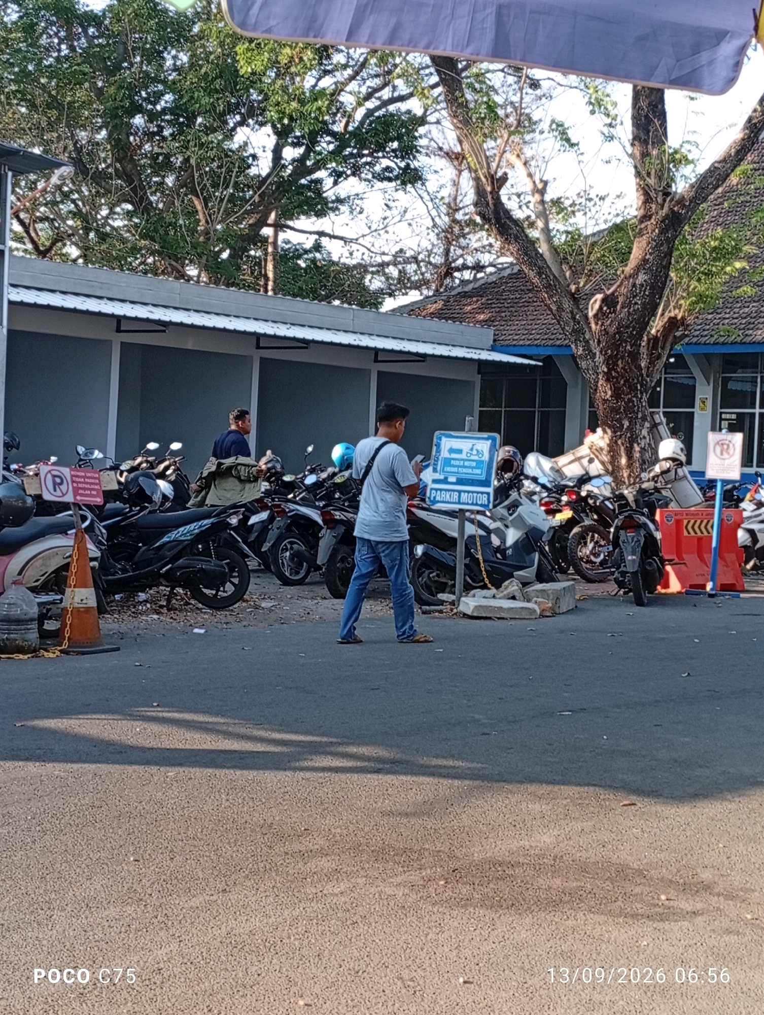 The image depicts an outdoor parking area with numerous motorcycles, suggesting it is a public space in Jepara, Indonesia. In the foreground, a man in a grey t-shirt and blue jeans is standing, looking at his phone. Behind him, a collection of parked motorcycles is visible, with a sign indicating "PARKIR MOTOR" (Motorcycle Parking). To the left of this sign, another sign with a "P" symbol inside a red circle is present, likely signifying parking restrictions. The environment includes trees with dense foliage, a building with a white roof and grey walls, and an asphalt ground surface. The lighting suggests it is daytime, possibly morning or late afternoon, given the shadows cast on the ground. A few traffic cones are also present in the area, one of which is orange and positioned near a chain. Another man in a dark shirt is seen walking away from the camera in the background, carrying a jacket over his shoulder. The overall scene suggests a typical day in a busy urban or institutional area in Jepara.