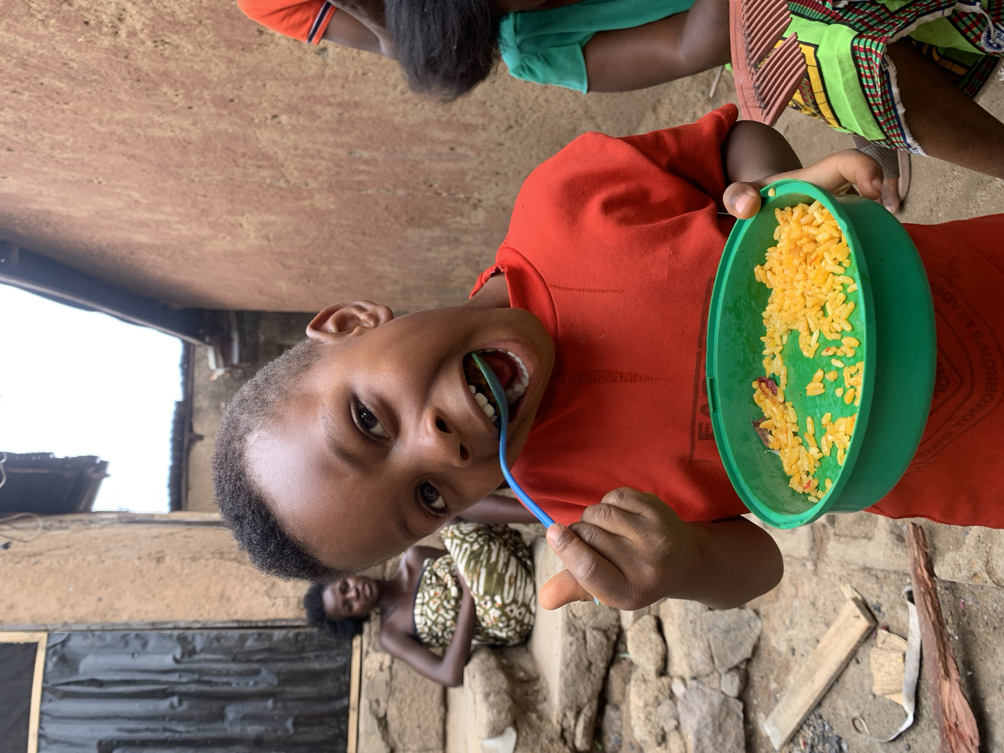 A young child, wearing a red shirt, is eating from a green bowl of yellow food with a blue spoon. The bowl is in their left hand, and the spoon is in their mouth. Behind the child, on the ground, a person lies wrapped in patterned fabric. The scene appears to be outside, possibly in Jos, Nigeria, given the description provided. Rough stone and wood debris are scattered around the ground, adjacent to a building with a dark metal sheet affixed to the side. Above and behind the child, other people's legs are visible, along with colorful patterned fabrics. The background includes walls and a glimpse of the sky.