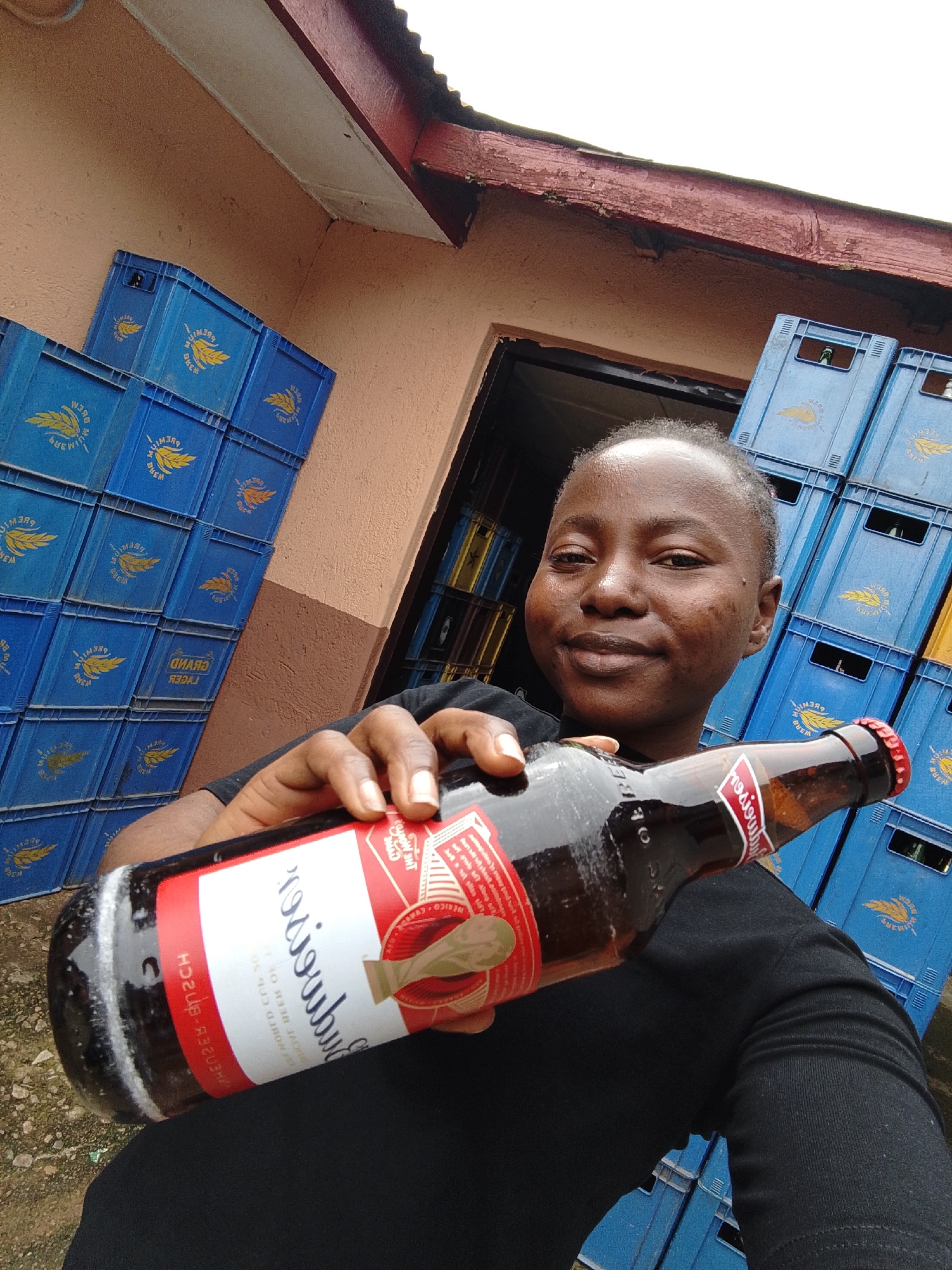 A young woman of African descent, with short hair and dark skin, is holding a large beer bottle in her right hand. She is wearing a black t-shirt. She is standing in front of stacks of blue plastic crates, some of which are filled with bottles and others appear empty. The crates are arranged in a way that suggests they are stored outdoors, possibly in a commercial area or a storage facility. The background shows a building with a textured peach-colored wall and a red corrugated metal roof. The overall impression is that of an outdoor storage area for beverages in an urban setting. The scene could be located in the city of Jos, Nigeria, known for its commercial activities and beverage distribution centers.