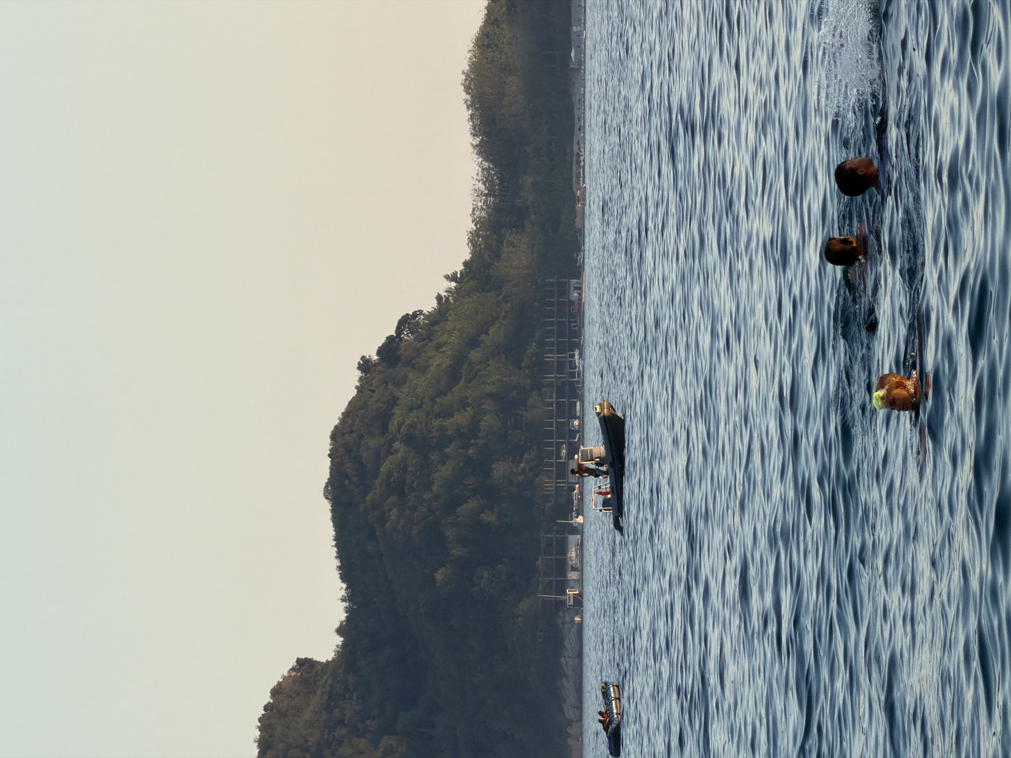 In the waters off Bečići, Montenegro, a scene unfolds with a lush, tree-covered hillside forming the backdrop to a calm sea. In the foreground, three individuals are swimming, their heads visible above the gentle ripples. One swimmer wears a yellow swim cap, and another is captured mid-splash, indicating activity. Further out on the water, a black inflatable raft carries at least two people. A larger, dark-colored boat with a person standing at the helm is positioned to the right of the inflatable raft. In the distance, along the shoreline, a partially constructed building with a visible framework stands near a line of beach chairs and umbrellas, suggesting a developing resort or tourist area. The sky above is clear and pale, indicative of late afternoon or early morning light.