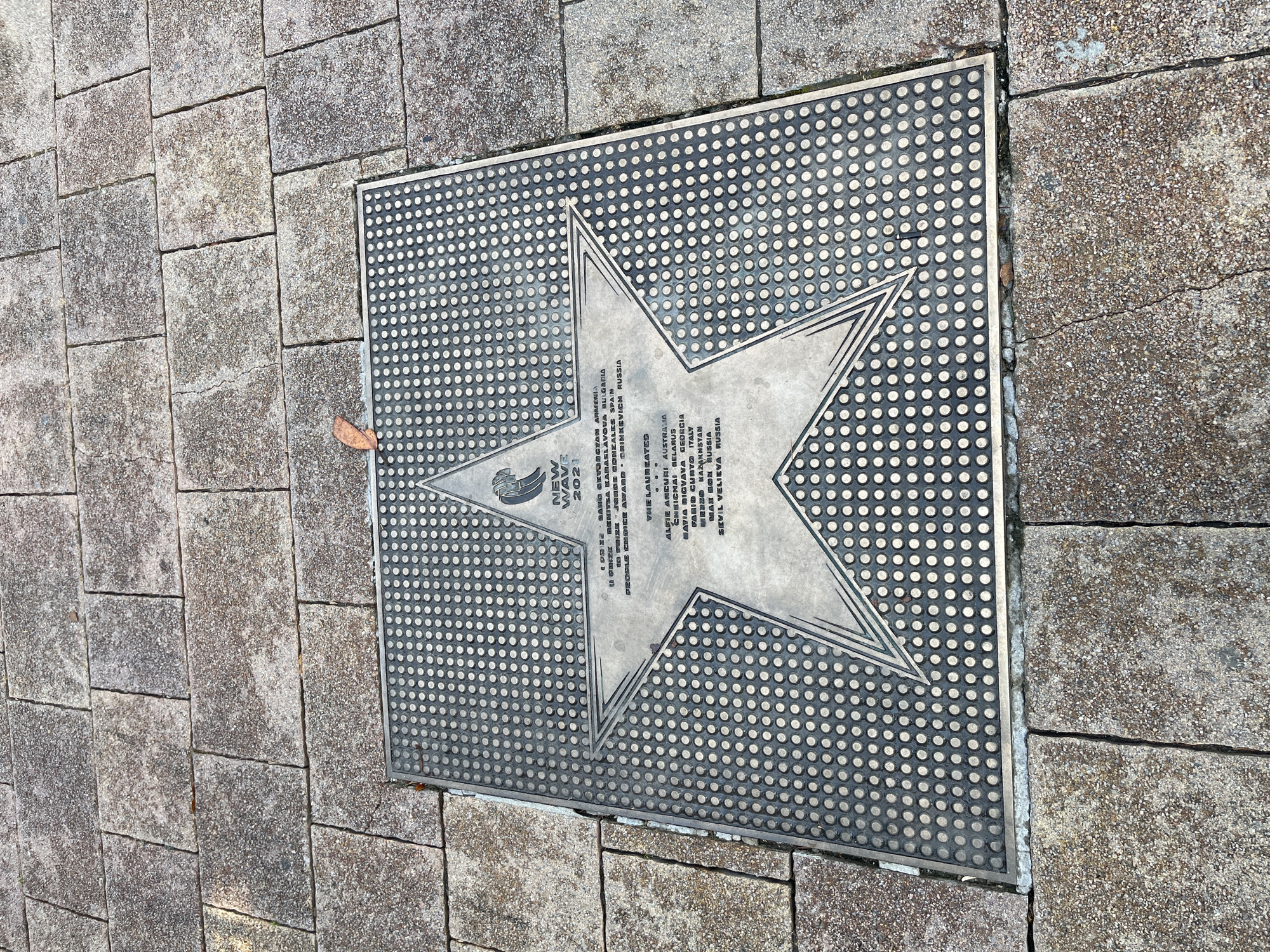 This is a photograph of a star plaque embedded in a paved walkway. The plaque is a star shape surrounded by a grid of small circles. The star has text inscribed on it, including the words "NEW WAVE 2021," which suggests it is related to an event. There is also text listing names and locations, such as "SANO GEVOEGYAN ARMENIA," "DENITSA KABASLAVOVA BULGARIA," and "GRINKEVICH RUSSIA," indicating this is likely a walk of fame or award for participants. The paving stones are square and rectangular, with visible cracks between them. A single dried leaf rests on the walkway near the plaque. The lighting suggests it is daytime, and the scene appears to be outdoors. The location context indicates this is in Sochi, Russia.