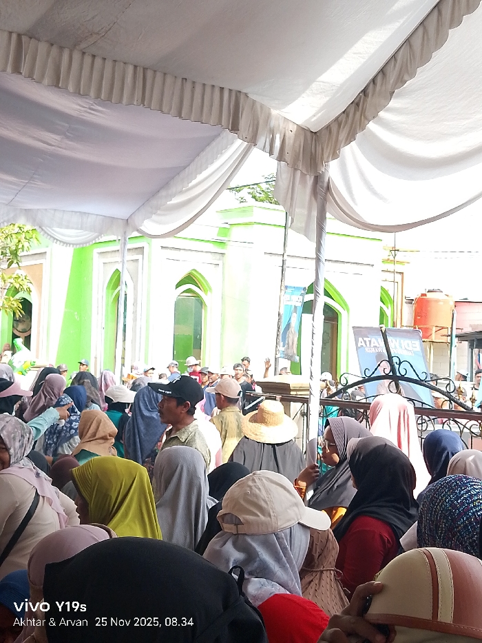A large crowd gathers under a white tent, with a green and white building visible in the background. Many people are wearing head coverings, including hijabs and hats. A few individuals stand out, one in a sunhat and another in a black baseball cap. In the background, a water tank and signs with text are visible. The image appears to be taken from within the crowd.