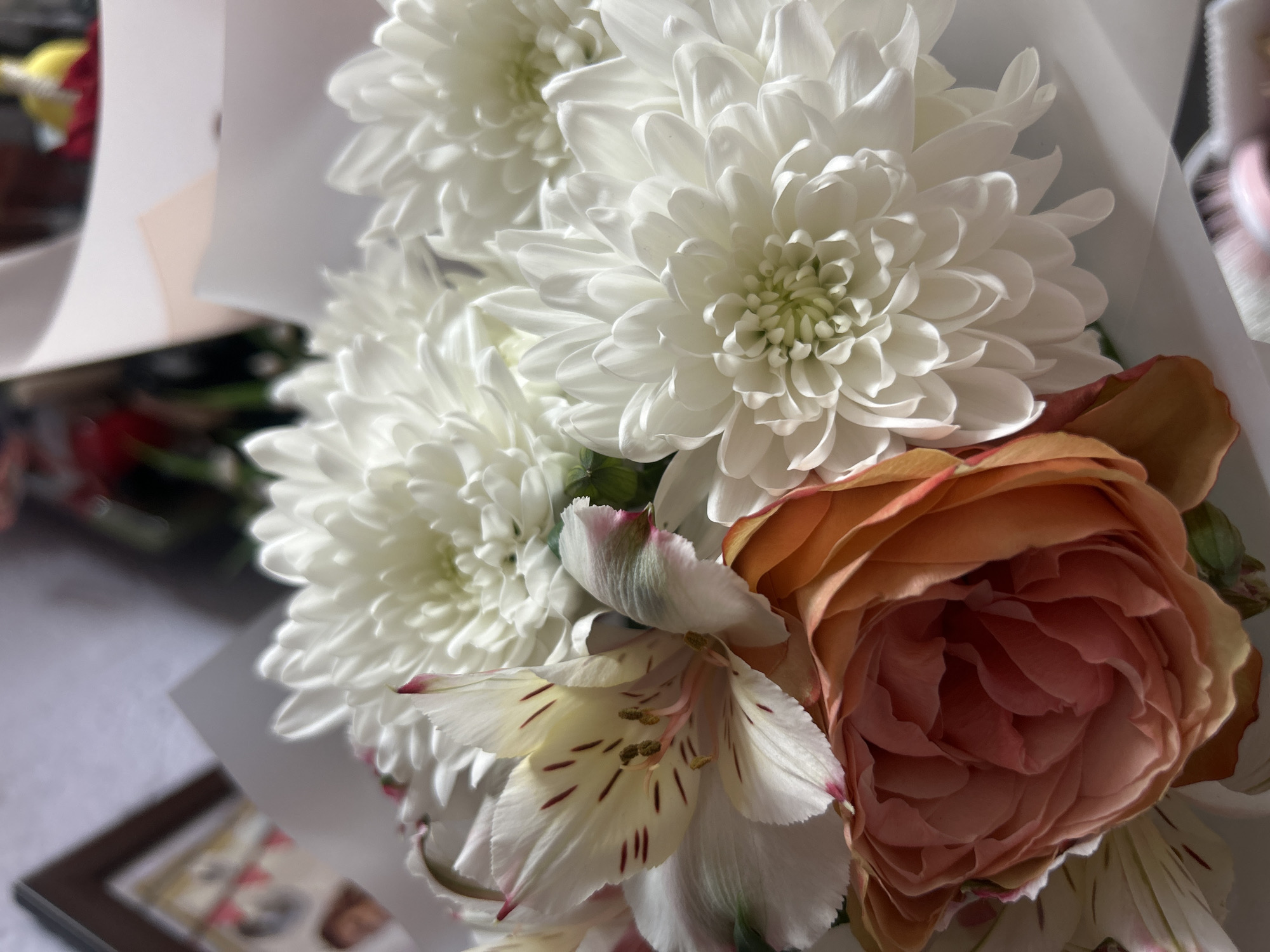 The image shows a bouquet of flowers. There are three white chrysanthemums, a pink and orange rose, and several white alstroemeria with red speckles. The flowers are arranged in a cluster, with the chrysanthemums in the background and the rose and alstroemeria in the foreground. The bouquet is wrapped in white paper. The background is blurred, but it appears to be a room with a picture frame on a table. Location: Unknown, Unknown.