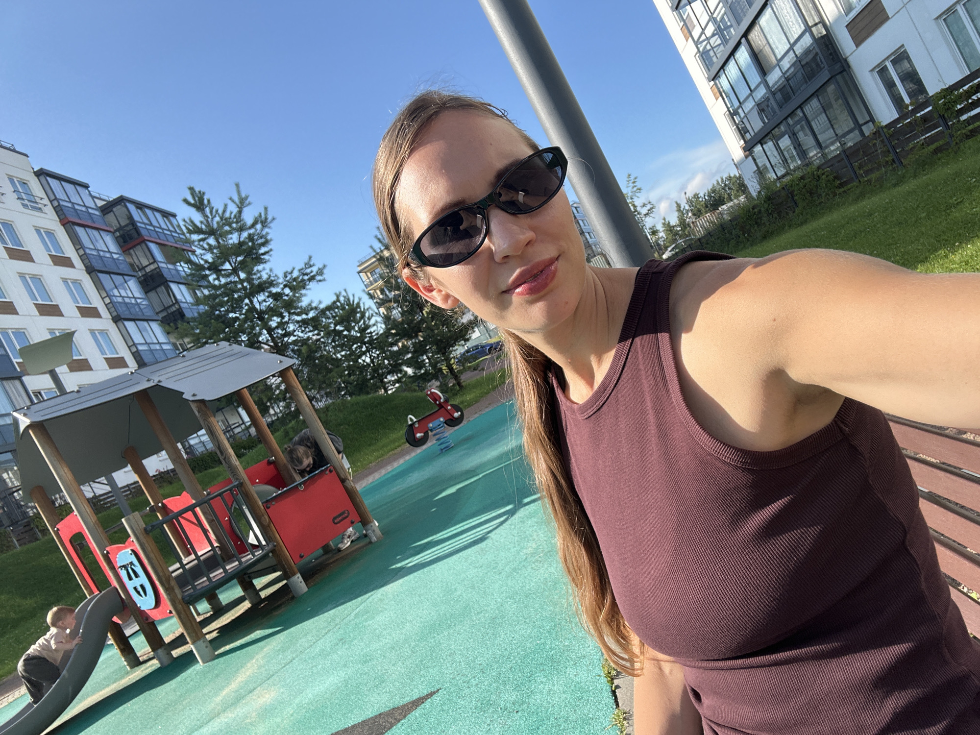 A woman wearing sunglasses and a dark tank top takes a selfie in a playground in Vsevolozhsk, Russia. The playground has green turf, a small red toy vehicle, and a wooden play structure. In the background, there are modern apartment buildings and trees under a clear blue sky.