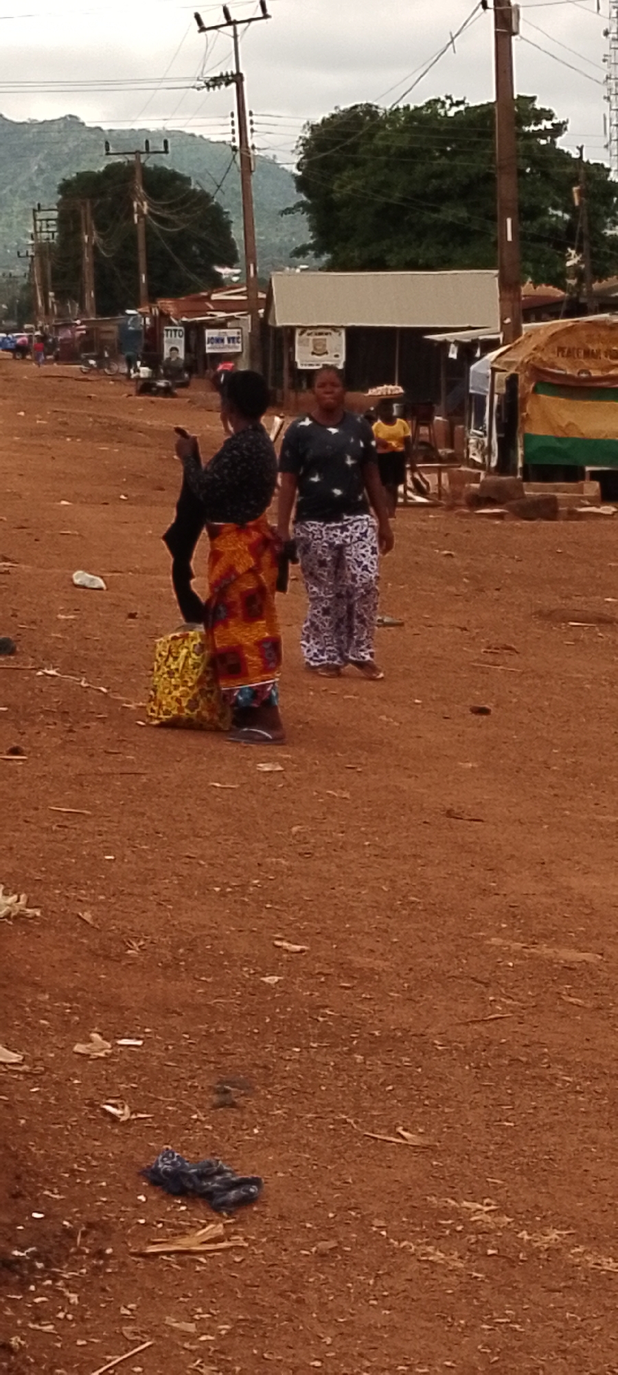 The image depicts an outdoor scene in Gboko, Nigeria. Two women are the central figures. The woman in the foreground is wearing a black patterned top and a vibrantly colored, patterned skirt. She is holding a dark garment in her left hand and standing next to a yellow and red patterned bag on the ground. To her right, a second woman, facing forward, is wearing a dark patterned t-shirt and patterned trousers. In the background, there are various structures that appear to be market stalls or shops, with signage visible on some. One sign reads "TITO" and another "JOHN VEC". There are also utility poles with overhead wires present. The ground is reddish-brown dirt, with some scattered debris. A pile of dark fabric is visible on the ground in the lower left of the frame. The sky is overcast. The overall impression is of a bustling outdoor market or street scene.