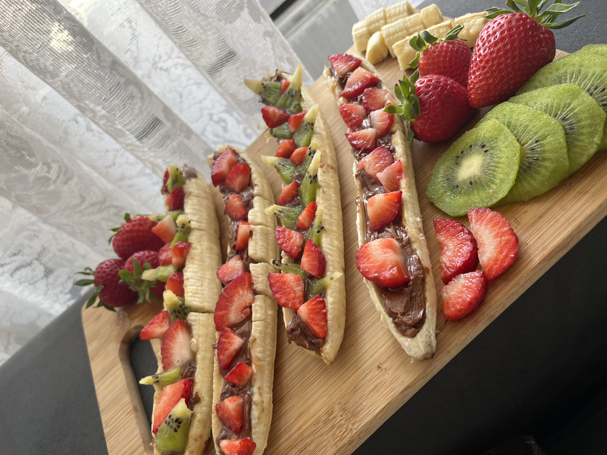 On a wooden cutting board, there is a display of sliced bananas filled with a dark spread and topped with chopped strawberries and kiwi. Several whole strawberries and sliced kiwis are grouped together next to the bananas, along with a row of sliced bananas. The cutting board is sitting on a dark countertop, and there is a white, patterned curtain in the background. The location is not discernible as being in Sofia, Bulgaria.