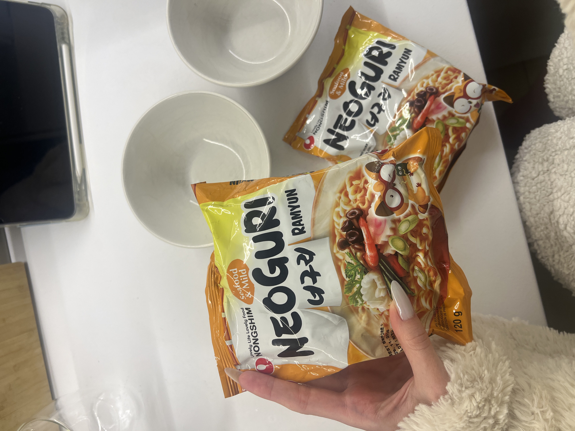 The image shows two packages of Nongshim Neoguri Seafood & Mild ramen noodles and two empty white bowls on a white table. A hand with long fingernails holds one of the ramen packages. A cell phone is visible in the upper left corner of the image, and part of a furry white garment is in the right side of the frame.