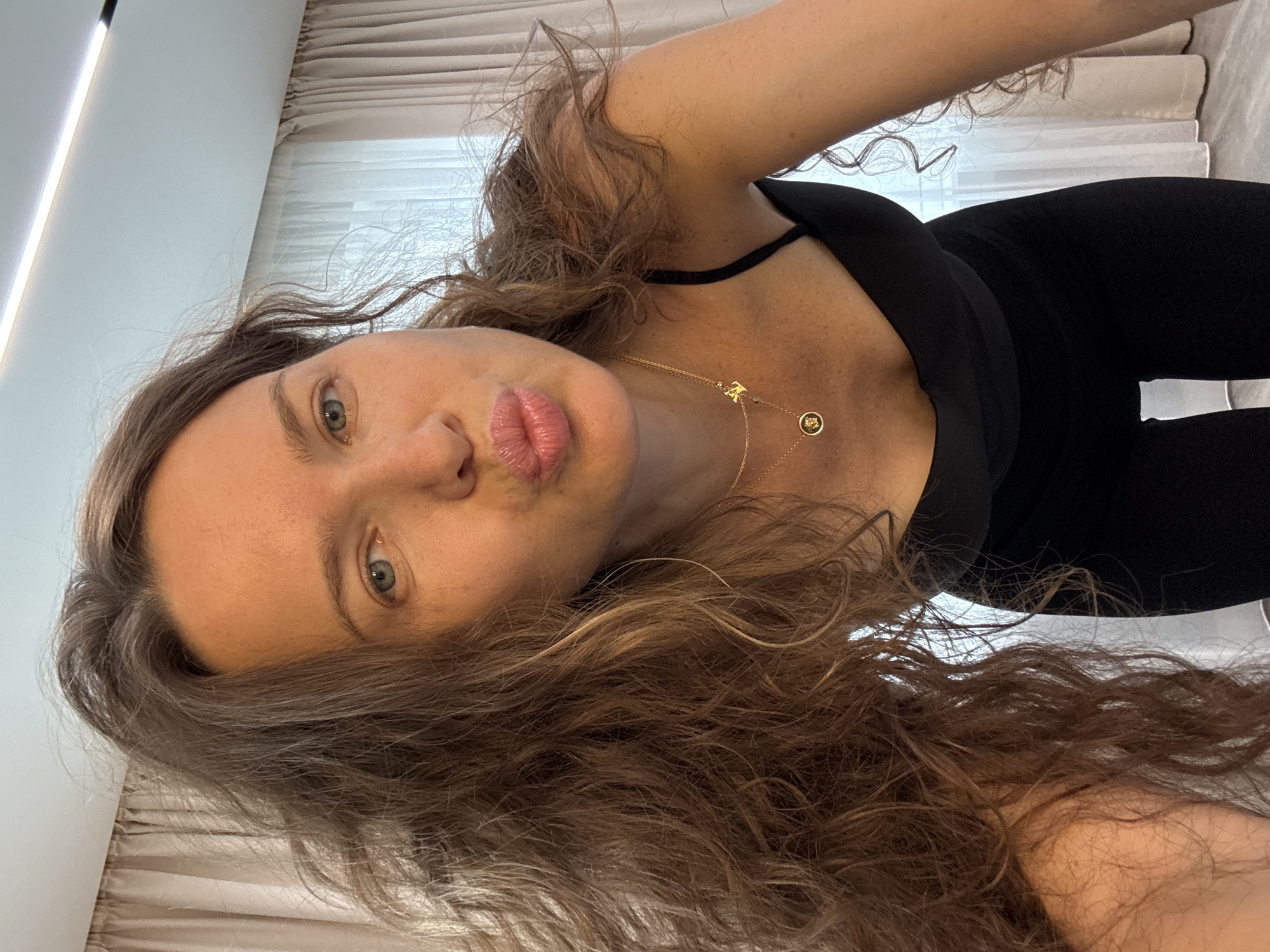 An adult female with long, wavy brown hair is looking into the camera with her lips pursed. She is wearing a black camisole and a gold necklace. The background consists of a sheer curtain and part of a ceiling light fixture. The scene is indoors, likely in a residential setting. No specific location in Khimki, Russia can be determined from the image.