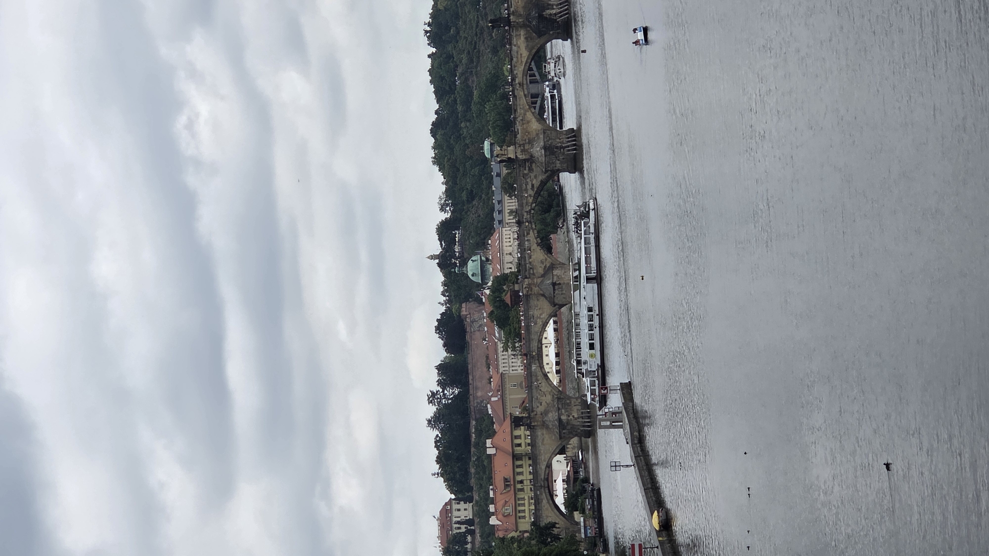 The image depicts a scene in Prague, Czechia, likely the Vltava River. A large, historic stone bridge with multiple arches spans the river. On the bridge, there are buildings with red-tiled roofs and visible people walking along it. A large white tour boat is moving on the river, parallel to the bridge. In the distance, a lush green hillside with trees and buildings, including a domed structure, rises behind the bridge. The sky is overcast with clouds. To the right, a small boat with at least two people is visible on the water. Several ducks are also present in the river. A pier or walkway extends from the left bank into the water.