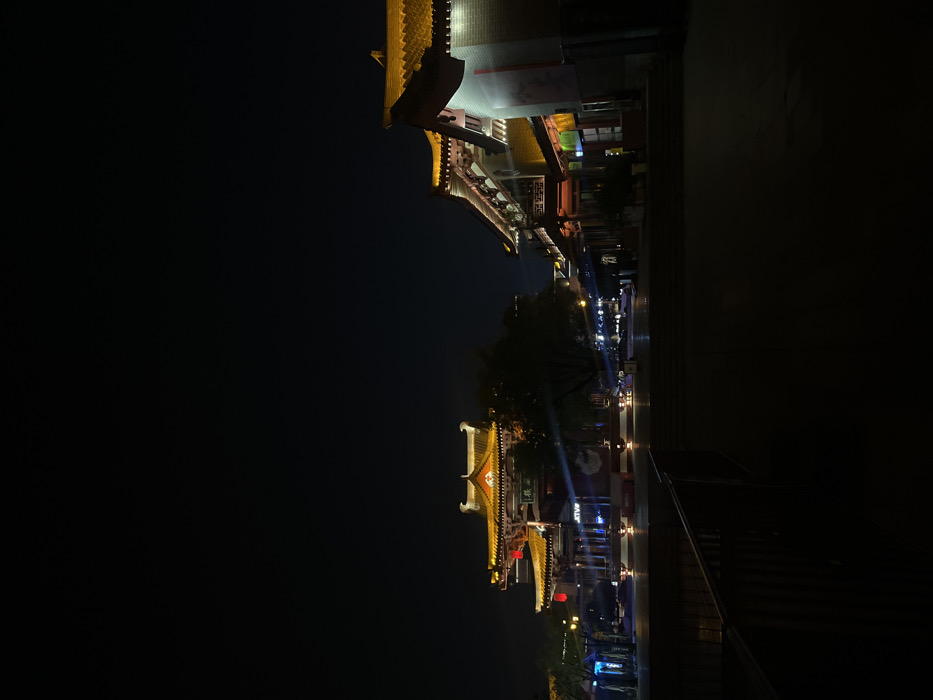 This nocturnal image depicts a traditional Chinese streetscape in Yanta District, China. The scene is dominated by multi-tiered buildings with ornate, upturned eaves, illuminated primarily by warm golden-yellow lights.
In the center-left, a prominent multi-story traditional Chinese pavilion stands, featuring intricate architectural details and multiple illuminated roofs. Red lanterns are visible hanging from its lower tier. A sign with Chinese characters is discernible on its facade. To the right, a row of interconnected traditional Chinese buildings extends along what appears to be a street or pedestrian pathway. These structures also exhibit illuminated golden-yellow roofs and some red accents. The ground in the mid-distance is an open, paved area, dimly lit by ambient sources and reflections. A dark metal railing is partially visible in the lower-left foreground, suggesting an elevated vantage point or a barrier alongside a lower level.
The sky is dark and clear, indicating nighttime. No identifiable individuals are present in the immediate foreground or mid-ground. Distant light sources and reflections are visible further down the street on the right. A prominent lens flare originates from a light source near the central pavilion. The overall environment appears quiet and meticulously maintained, highlighting the architectural elements through strategic illumination.