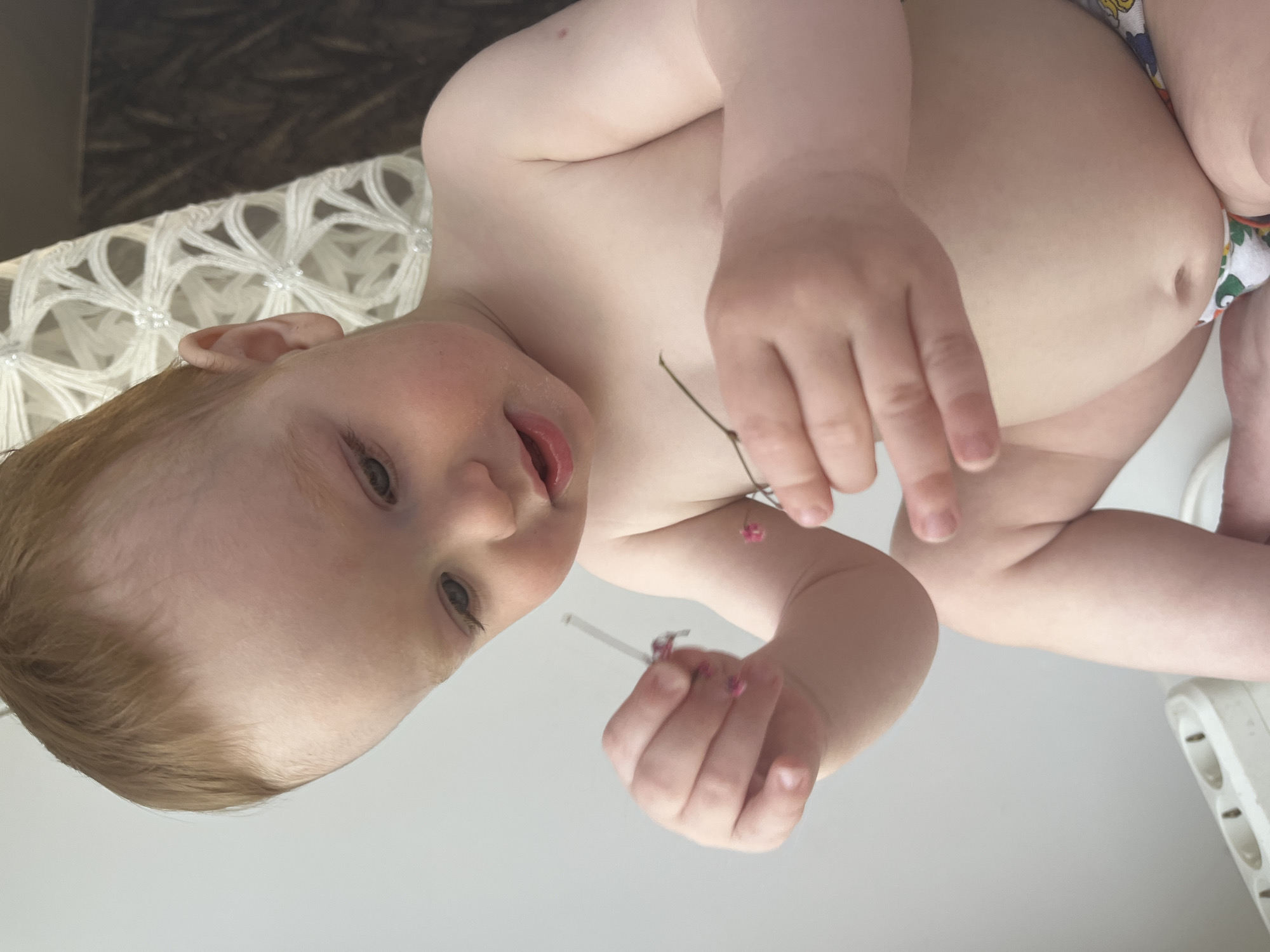 A fair-skinned infant is shown from an overhead perspective, with their head toward the left of the frame. They have red hair and are looking at the camera with a slight smile. The child holds a thin plant stem with small pink flowers in their hands. They are wearing a diaper with a colorful print. The background includes a white surface with a textured, possibly crocheted edge at the upper left, a dark mottled surface, and a white multi-socket power outlet visible at the lower right. The photograph was likely taken indoors in the city of Chaikovsky, Russia.