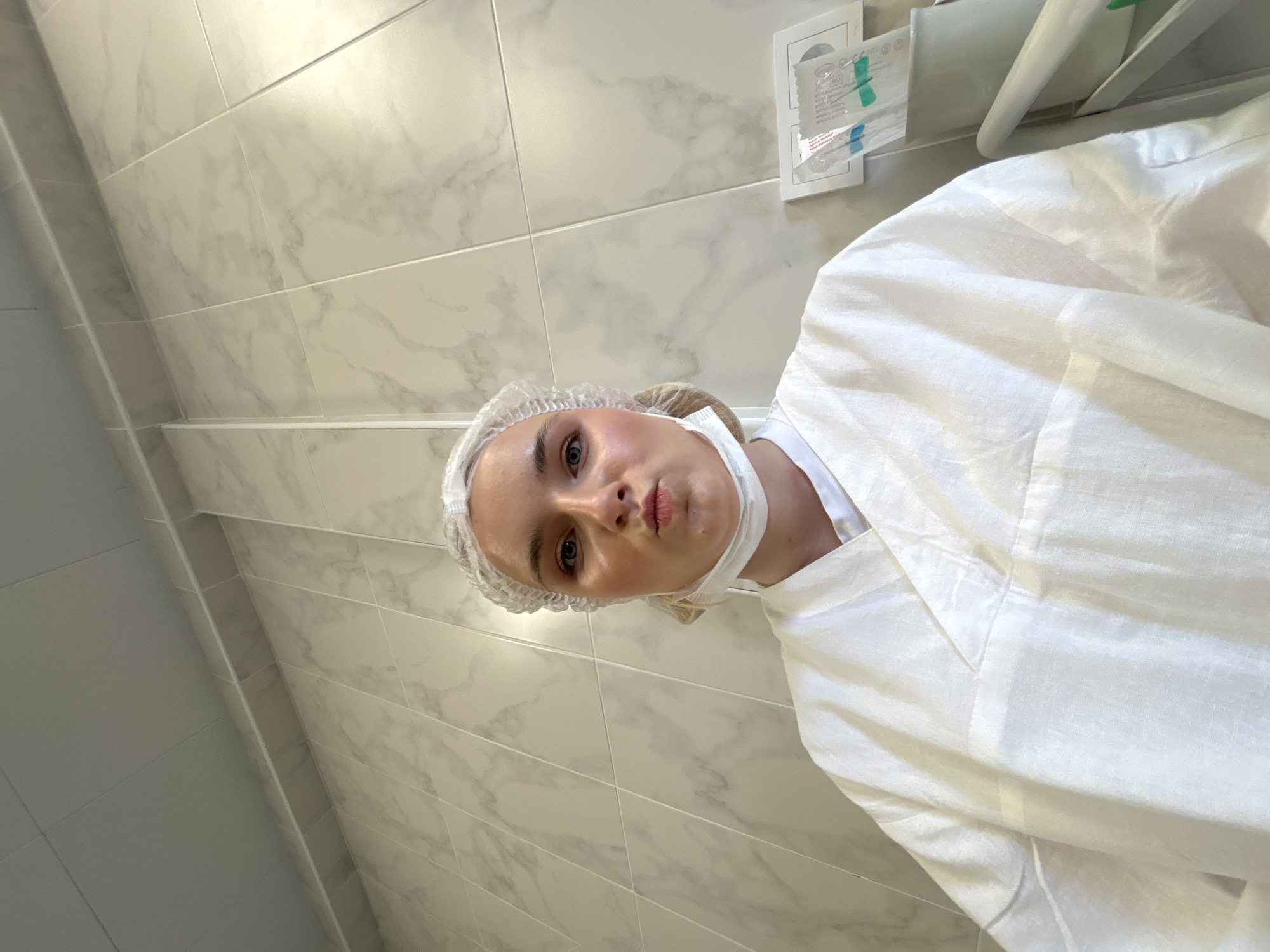 A person with fair skin is shown from a low-angle perspective, wearing a white surgical cap and a white medical gown. A white surgical mask is loosely positioned around their neck. The person has light-colored hair and is looking directly at the camera with a slight pucker of the lips.
The background consists of white marble-patterned tiled walls. Near the upper-right corner, there is a white medical device or fixture mounted on the wall, with disposable items attached.
There are no indications that the image was taken in Saint Petersburg, Russia.