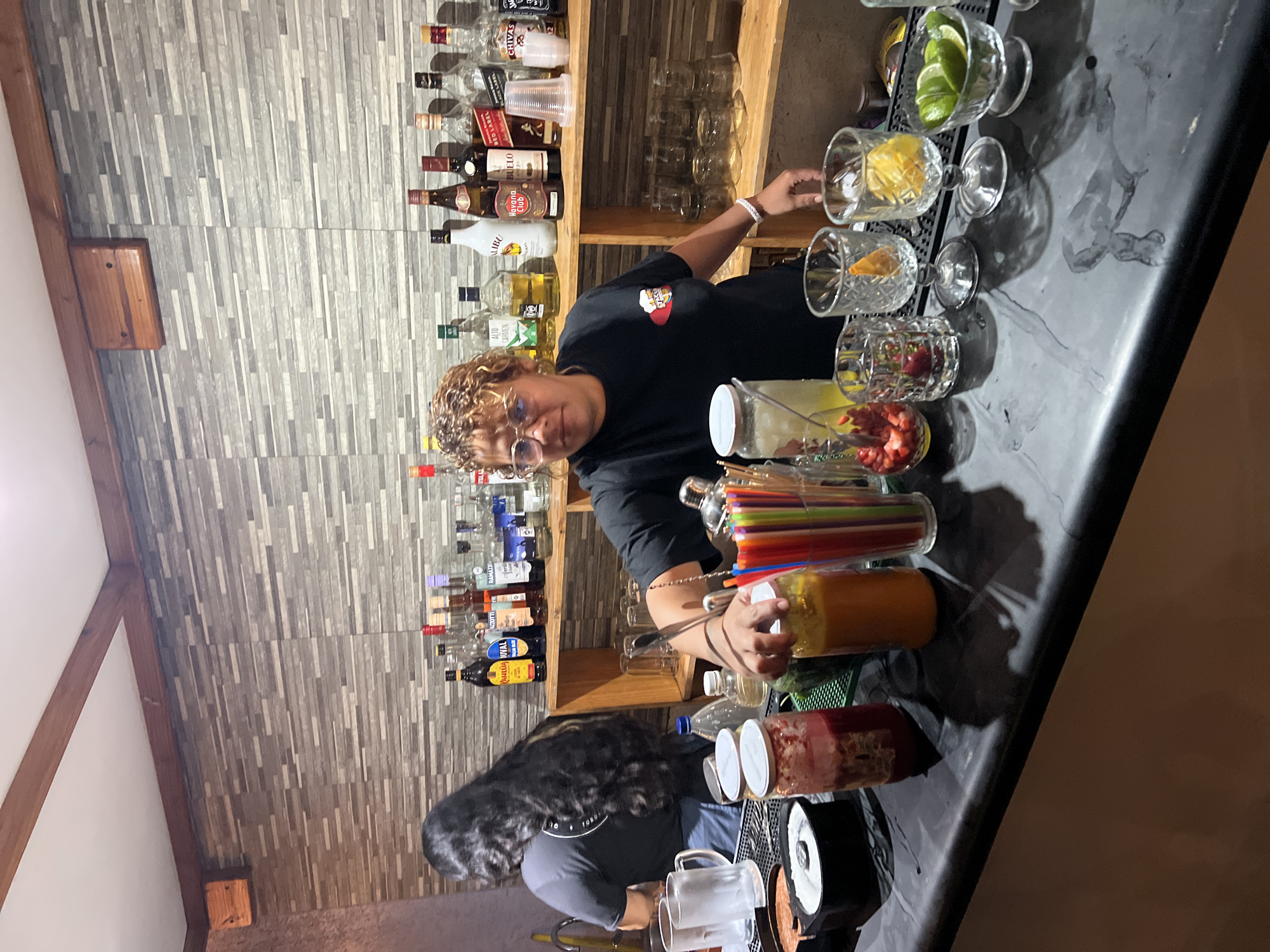 A photograph shows two people behind a bar in what appears to be Arica, Chile. One person, an adult, has curly hair and wears glasses and a black shirt with a small graphic on the chest. They are positioned behind a bar containing various liquor bottles and glassware. The other person has long dark hair, and wears a light colored shirt. A variety of cocktail preparations sit on the bar, including cut fruit and juices. Glass jars hold colorful straws and other garnishes. The bar itself is dark, possibly made of granite or a similar material. The wall behind the bar is decorated with a grey striped pattern. The lighting is dim, giving the image a somewhat warm tone. The overall setting indicates a casual, possibly festive atmosphere.