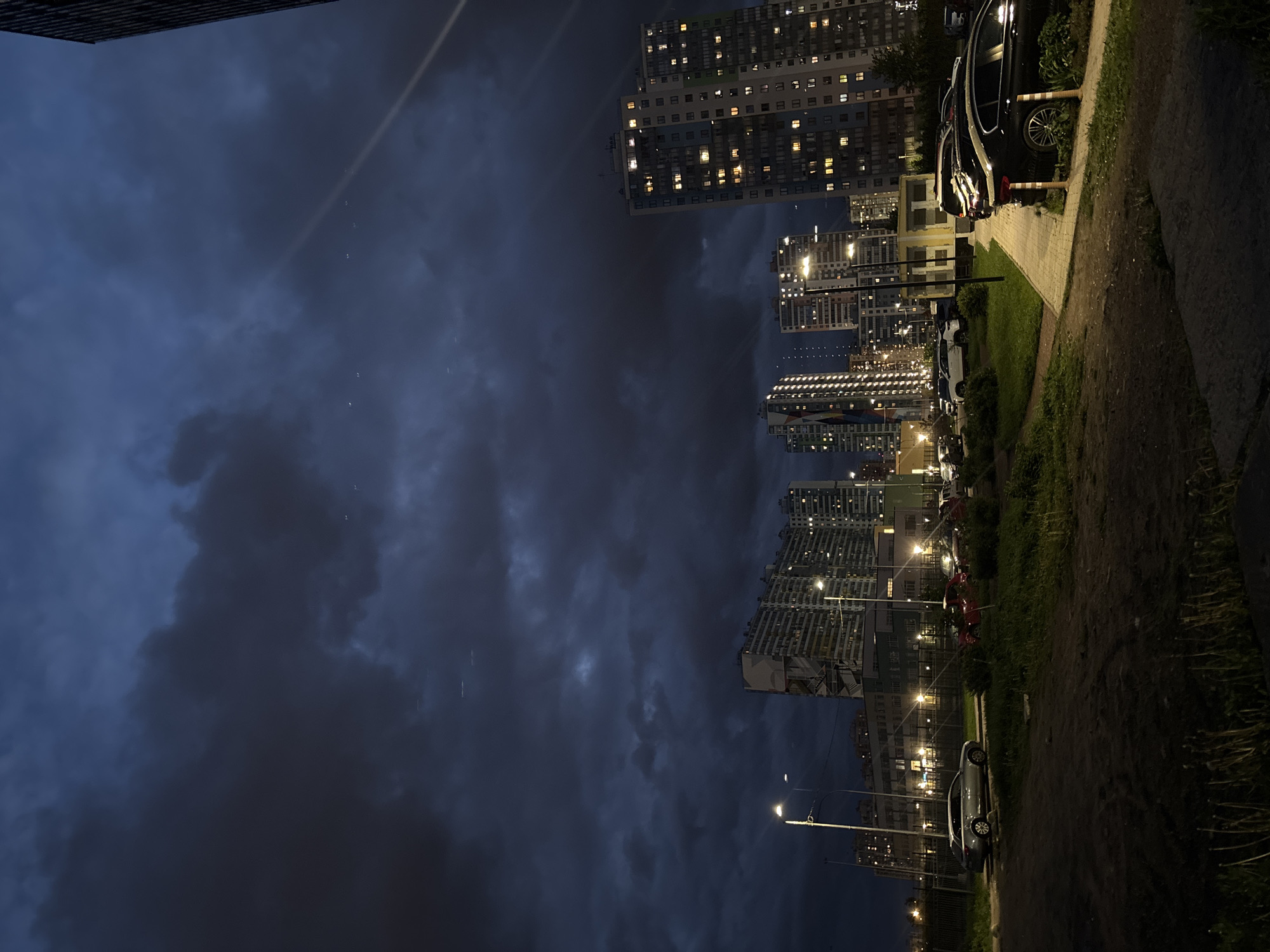 The image depicts a night scene in a city that appears to be Saint Petersburg, Russia. The perspective is unusual, as the photo is rotated and taken at a low angle. In the foreground is a dark asphalt pathway bordered by grassy areas. Several cars are parked along the edge of the road. Streetlights illuminate the scene, casting a warm glow. Tall apartment buildings are visible in the background, with many windows lit up, indicating activity inside. The sky is filled with dark, heavy clouds, possibly indicating a storm.