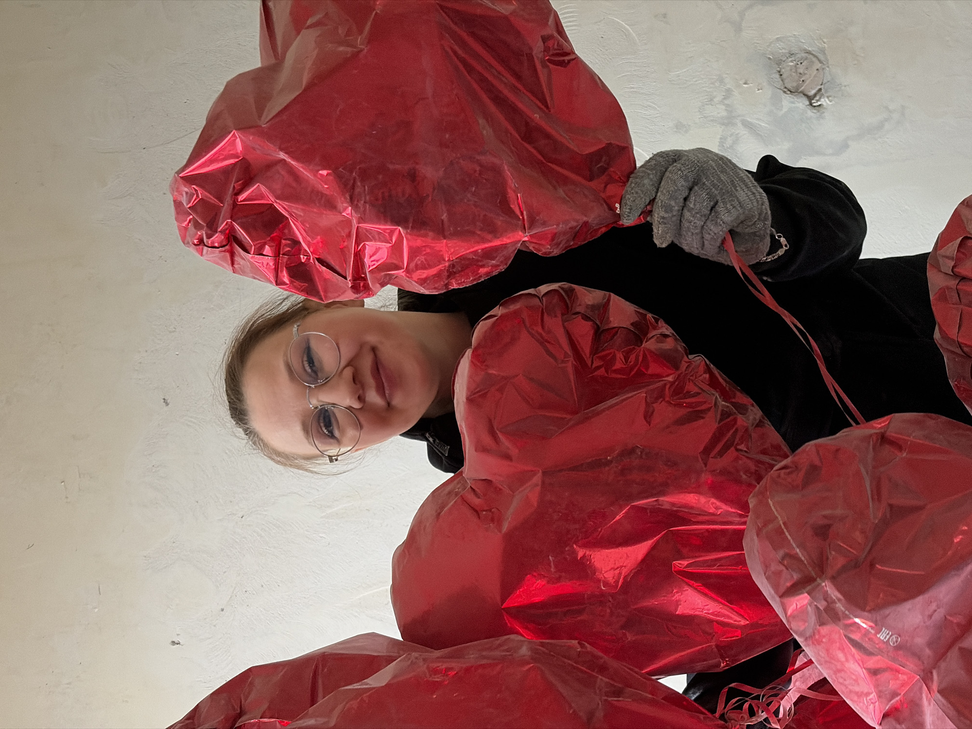 An upward-angle shot captures a young woman holding multiple red, heart-shaped foil balloons in an indoor setting in Izhevsk, Russia. The woman is visible from the chest up, with light-colored hair, round metal-rimmed glasses, and a soft, pleasant smile directed towards the viewer. She is dressed in a dark (likely black) hooded top, and her right hand, clad in a grey knitted glove, holds a red ribbon connected to a balloon. A silver chain is visible on her right wrist.
The foreground and midground are dominated by the shiny, metallic red balloons, some appearing slightly deflated or creased. The background is a textured, light-colored wall or ceiling, possibly concrete or plaster, showing various imperfections, discolored areas, and a small, rough patch near the top right. Faint, white text resembling "EAC" and a recycling symbol can be seen on the surface of one of the balloons in the lower right. The image conveys a cheerful or celebratory mood, likely associated with an event such as Valentine's Day or a birthday. The time of day is not indicated, and the lighting appears to be indoor.