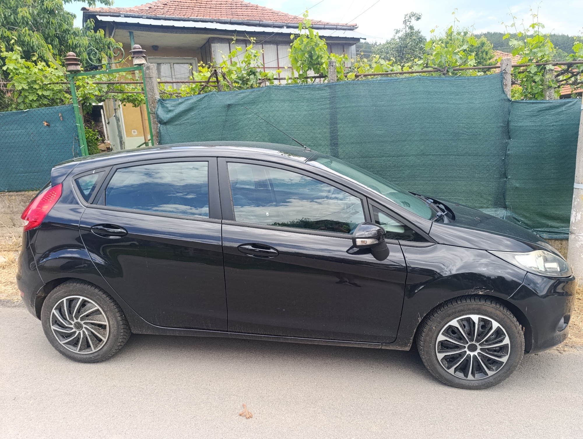 A black Ford Fiesta hatchback is parked on the asphalt side of a road, facing right. The car shows some dust and dirt on its lower body panels and wheels. Notably, the front and rear wheels have different designs of wheel covers. There are minor scuffs on the black paintwork, particularly visible on the front fender above the right wheel. Reflections of clouds and sky are visible in the car's windows. An antenna extends from the car's roof.
The background features an older house with a reddish-brown tiled roof, partially obscured by lush green foliage, including grapevines, suggesting a garden or vineyard setting. A green privacy netting stretches along a fence made of concrete posts and horizontal metal bars, separating the road from the property. In the left portion of the image, an old, decorative metal gate, also overgrown with vines, is visible, topped with two distinctive ceramic electrical insulators. Behind the house and vegetation, distant hills covered with trees can be seen under a partly cloudy sky. The scene appears to be set during daytime in a rural or semi-rural area, consistent with a village like Nikolichevtsi, Bulgaria. No discernible text is clearly visible in the image.
