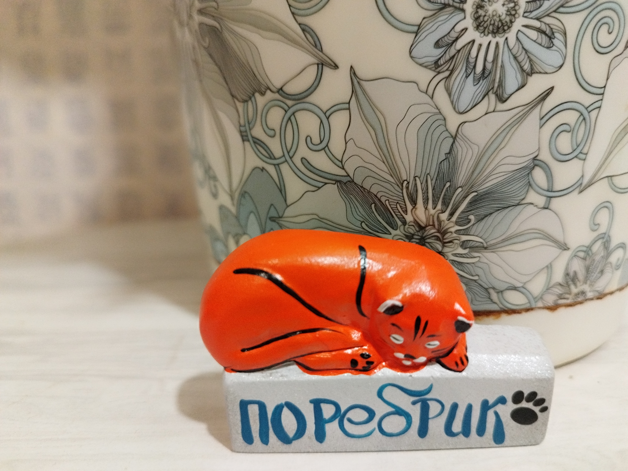 This is a close-up, eye-level shot of a small, orange, ceramic cat figurine sleeping on a white rectangular base. The base has blue Cyrillic letters painted on it, reading "ПОребиРk" and a black paw print. The cat is curled up with its tail tucked, and has black stripes on its back and face, with white accents for its eyes and whiskers. Behind the figurine, a portion of a ceramic pot with a floral pattern in shades of blue, grey, and white is visible. The surface the figurine rests on appears to be a light-colored wooden table. The image is well-lit, with soft shadows cast by the figurine. There is no indication of the city of Saint Petersburg, Russia in this image.