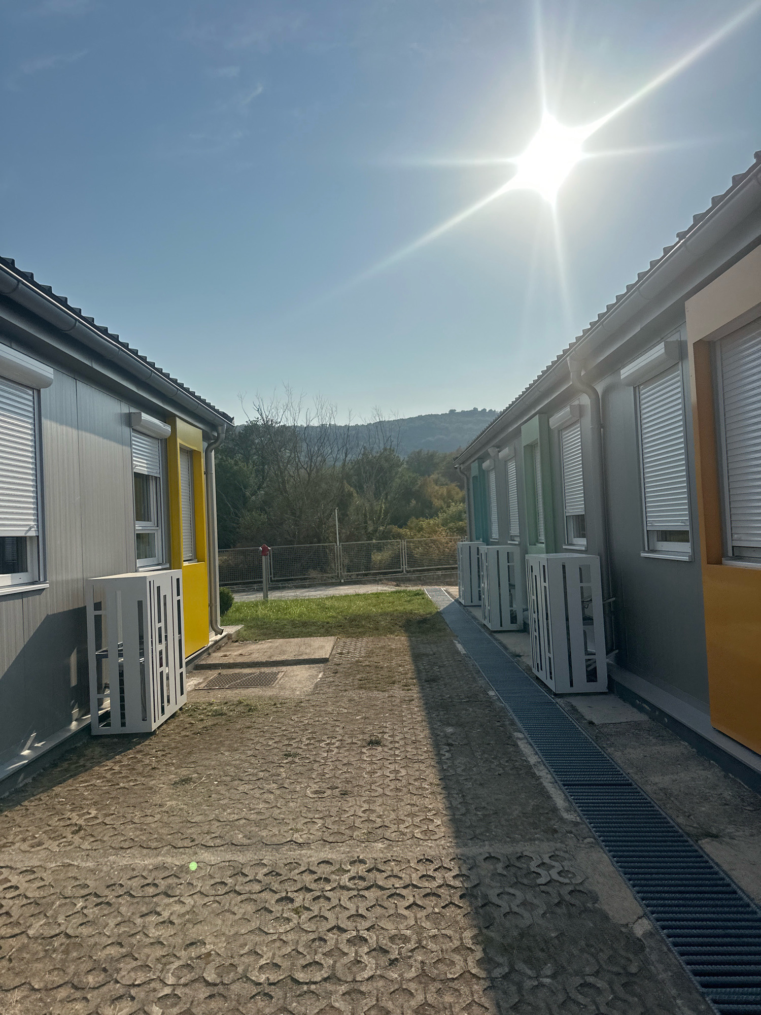 The image depicts an outdoor scene with two parallel rows of modular buildings. The buildings are gray with yellow and green accents and feature roll-down shutters over the windows. In front of each building are outdoor air conditioning units, enclosed in white decorative cages. The ground is paved with interlocking concrete blocks and a metal grate drain runs along the side of the right building. In the background, there is a grassy area, trees, and a distant hill under a bright, sunny sky with a lens flare from the sun. A red fire hydrant is visible near the fence in the middle ground. The location is identified as Radanovići, Montenegro.
