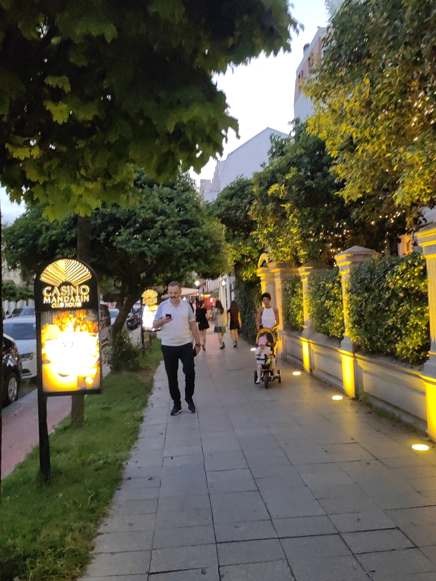 This media file shows a street scene with several people walking on a sidewalk. The time of day appears to be late afternoon or early evening, as the sky is a soft grey and artificial lights are on. The weather appears to be mild.
In the foreground, a man in a white polo shirt and dark pants is walking and looking at his phone. To his left is a brightly lit sign that reads "CASINO MANDARIN CLUB HOUSE" with an image of flames. Behind him, other people are visible walking along the sidewalk. Further down the sidewalk, a woman is pushing a child in a stroller. The sidewalk is paved with square tiles and lined with lush green bushes and trees. The bushes are illuminated by yellow lights, creating a warm glow.
The scene appears to be a bustling urban or tourist area, possibly a promenade or a street lined with shops and entertainment venues. The overall mood seems relaxed and casual. The lighting suggests the transition from day to night, and the presence of illuminated signs and pathway lights indicates an area that is active after dark. The architecture visible in the background, including buildings with multiple stories, contributes to the urban setting.