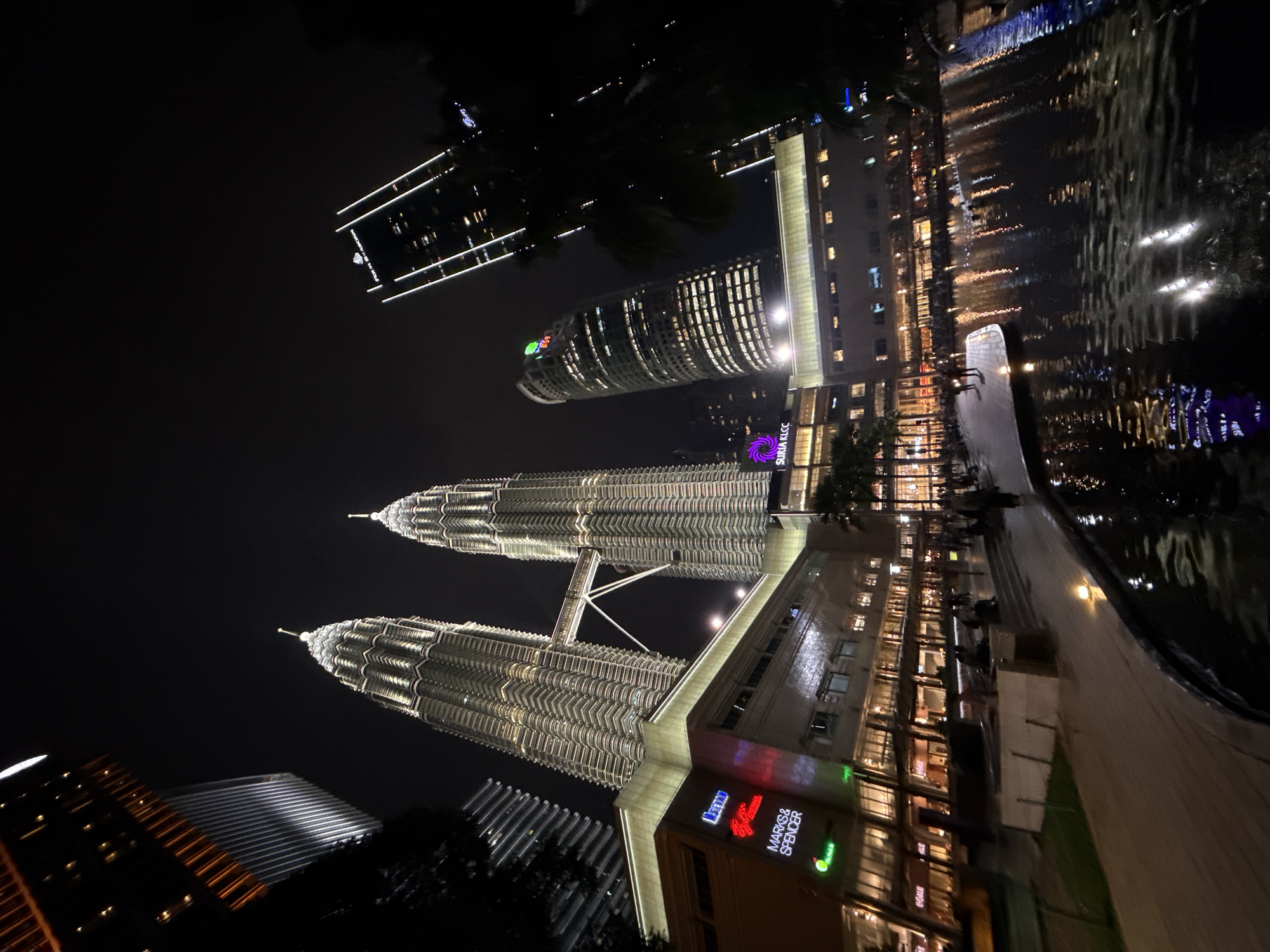 The image shows the Petronas Twin Towers in Kuala Lumpur, Malaysia, at night. The towers are illuminated, and their reflection is visible in a body of water. Other illuminated buildings are also visible in the background. The lower part of the image shows the Suria KLCC shopping mall. The area appears to be a walkway or plaza, possibly with people present, although they are not clearly visible. The sky is dark, suggesting it is nighttime.