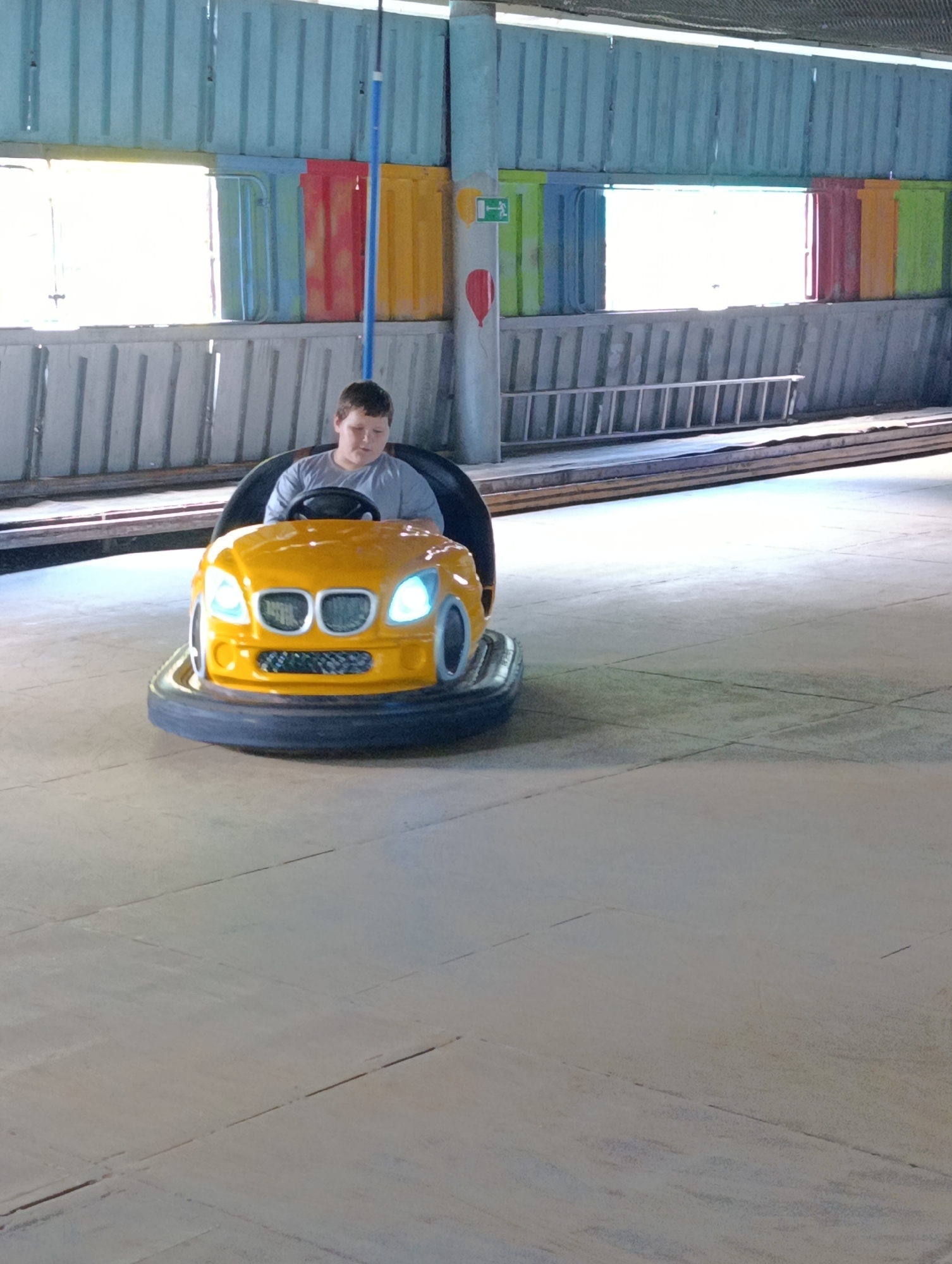 A young boy is seen driving a yellow bumper car. The bumper car is circular with black rubber bumper guards and white headlights. The boy is wearing a grey t-shirt and is looking down. He is seated in a darkened interior of the bumper car, with his hands on the steering wheel. The background shows a brightly lit area with a light blue structure above. The floor is a light grey, concrete surface. The image is taken from a medium-low angle, with the bumper car in the foreground and the background slightly out of focus. There is a ladder visible in the background on the right side of the image, and colorful panels are stacked behind it. The overall scene appears to be an indoor amusement area.