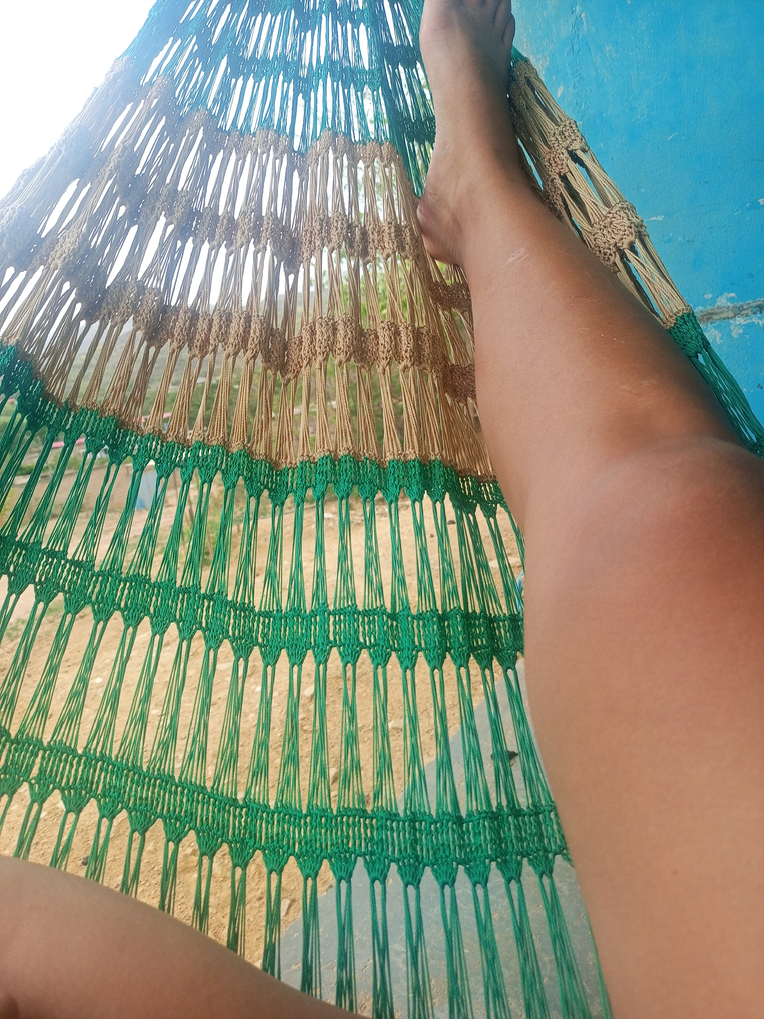 A person's legs are visible, resting in a hammock. The hammock is made of woven cords in shades of green, tan, and blue. Through the weave, a sandy ground and some vegetation are visible. The background is a bright blue wall, suggesting an outdoor setting. The lighting indicates it is daytime, possibly late afternoon given the angle of the sun creating some lens flare. The overall mood is relaxed and peaceful, suggesting a moment of leisure. There is no visible text in the image. The scene is set in La Tagua, Venezuela.