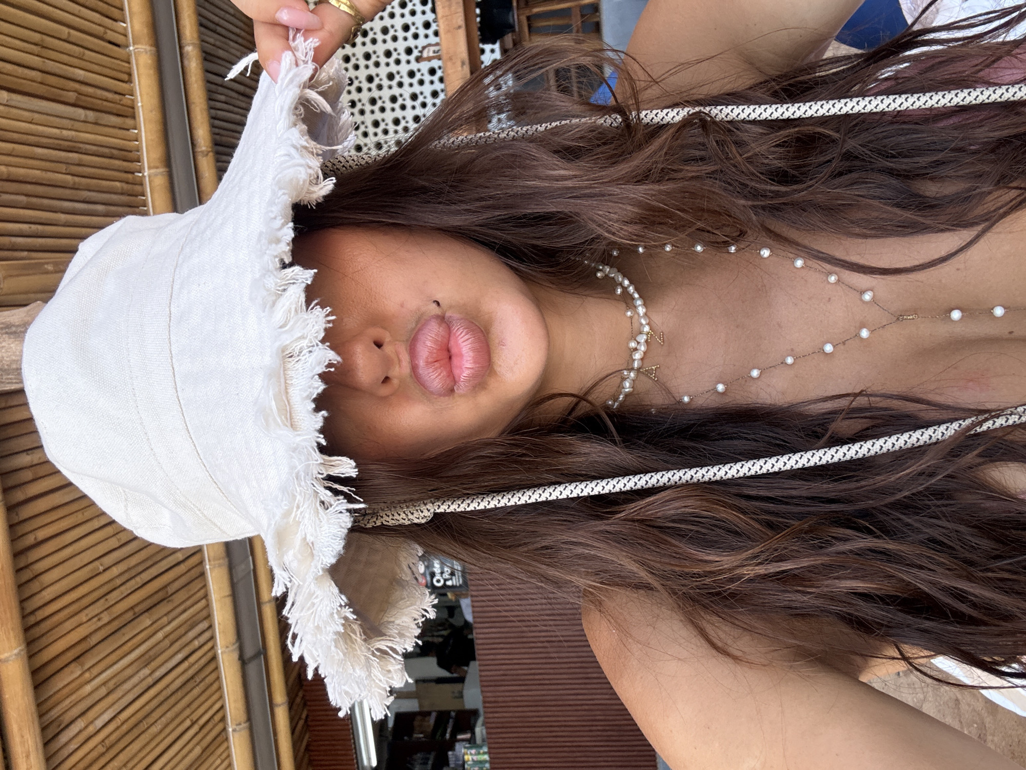 The image is a low-angle selfie of a woman with long, wavy brown hair. She is wearing a white bucket hat with a fringed brim. A white beaded necklace is visible around her neck, and a white rope-like head band is on her hair. Her lips are pursed. The background shows a bamboo structure and glimpses of an interior space with shelves or objects. The location of the photograph cannot be determined using the information available.