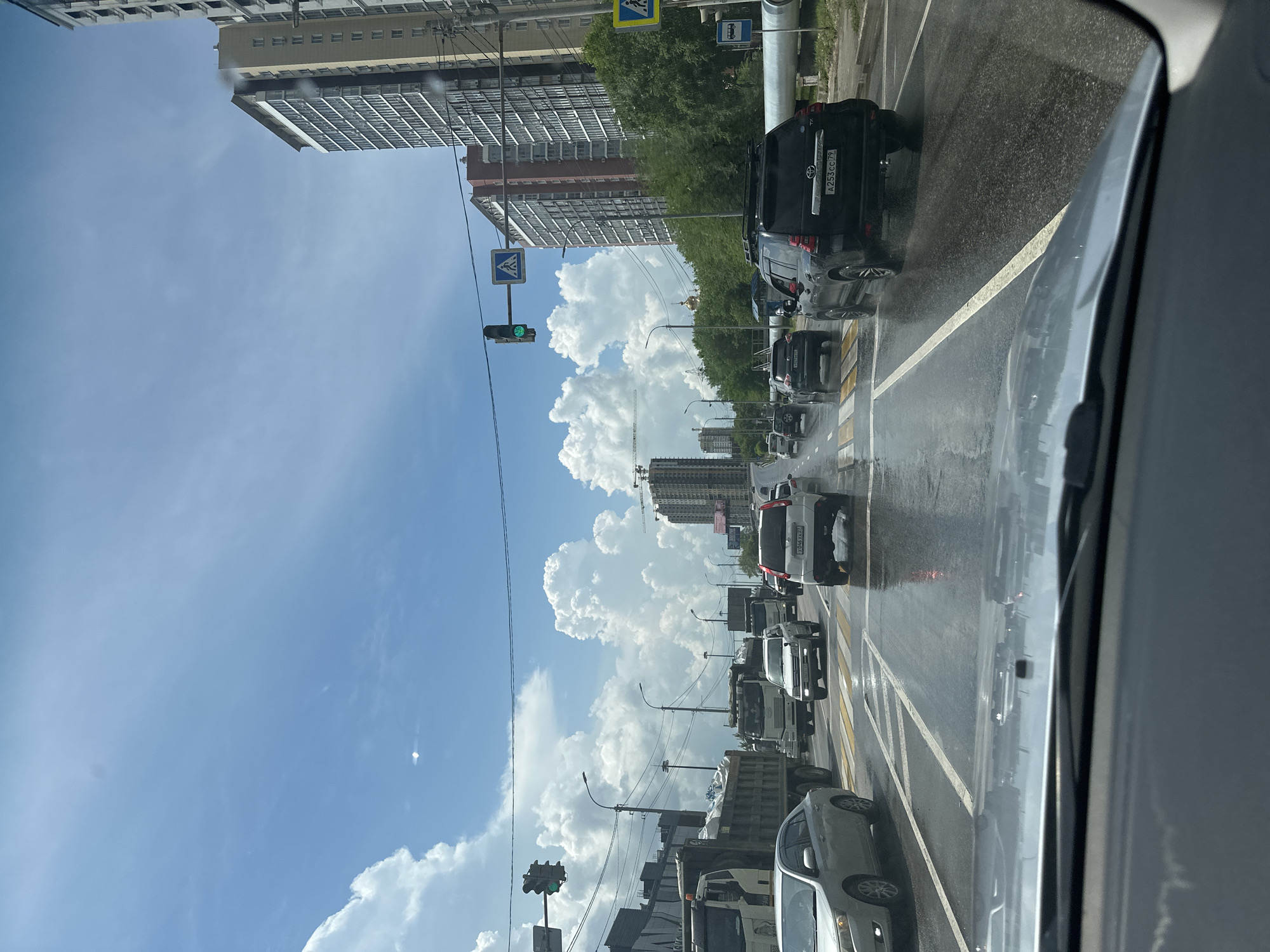 The image depicts a street scene in the city of Khabarovsk, Russia, likely on a cloudy day with the sun occasionally breaking through. The perspective is from inside a vehicle looking forward through the windshield. The road surface is wet, suggesting recent rain or ongoing precipitation. Traffic is moderate, with several cars, SUVs, and trucks visible on a multi-lane road.
In the foreground, the dashboard of the vehicle is partially visible. The road features clear lane markings and a pedestrian crossing. A traffic light is visible on the left, showing a green signal. Several large, modern apartment buildings are prominent along the right side of the road, indicating an urban setting. Trees line the median or sidewalk area, and construction cranes are visible in the distance, suggesting ongoing development. The sky is a mix of blue and white clouds, with some atmospheric haze. No people are clearly discernible.