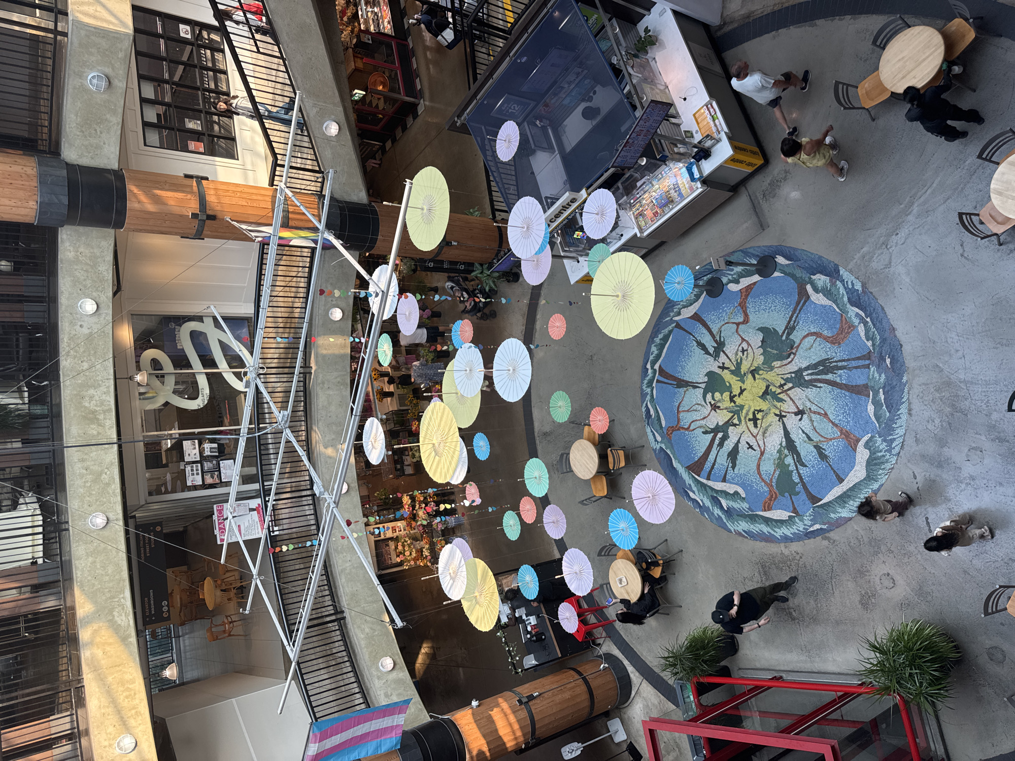 This is an aerial view of the interior of Lonsdale Quay Market in North Vancouver, Canada, on a sunny day. The market is a multi-level indoor space with a central atrium. Suspended from the ceiling are numerous colorful paper umbrellas in various shades of yellow, white, blue, pink, and green, creating a festive canopy. On the ground floor, a large circular mosaic artwork depicts a natural scene with trees, birds, and water. Several people are visible on the ground floor. A man in a black t-shirt and dark pants is walking towards the left. A woman is standing near a round table, appearing to be looking at her phone. Another group of people are near a counter with a refrigerated display, likely a food vendor.
Further up, on the second level, people are seen walking on a balcony overlooking the atrium. A large wooden pillar is a prominent architectural feature in the center of the image. Shops and seating areas are visible on the different levels, and stairs connect the floors. The overall atmosphere appears to be lively and commercial.