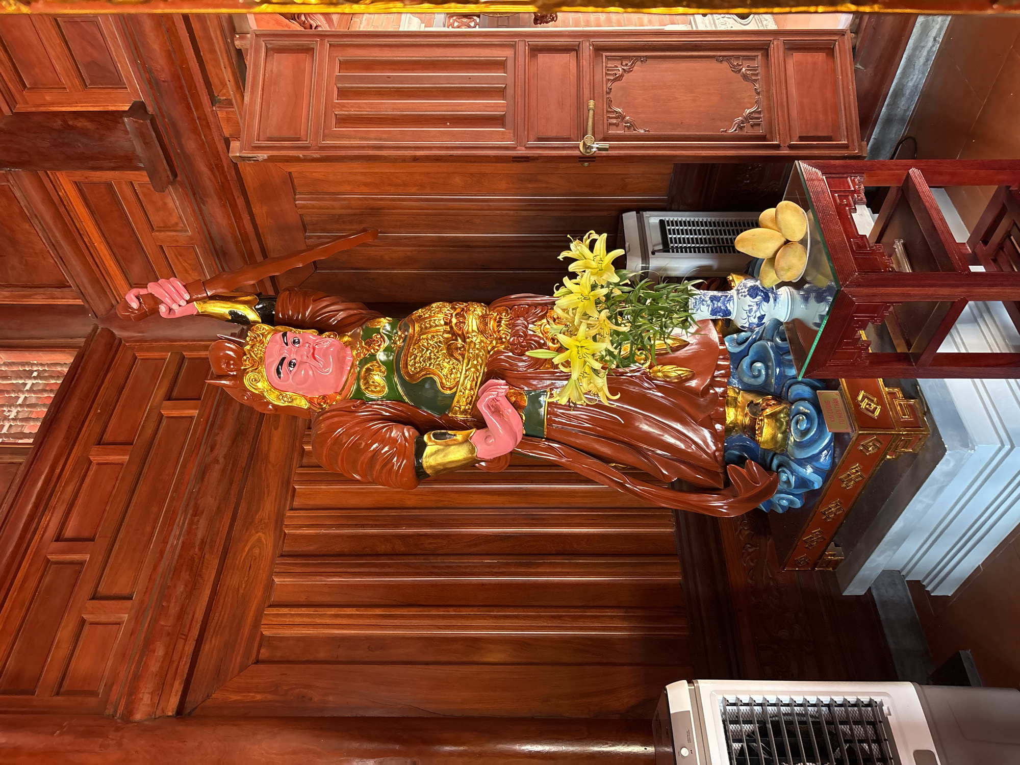 A statue of a deity stands on a table laden with offerings in what appears to be a temple or shrine in Nam Nha Trang Ward, Vietnam. The statue is adorned in a golden robe with red sleeves and an elaborate green collar, holding a sword in its right hand. The deity's face is painted pink. Yellow lilies and a blue-and-white vase are positioned in front of the statue. A plate of mangoes is located near the statue. An air purifier is seen beside the table. The surrounding environment features dark, polished wooden walls and furnishings.