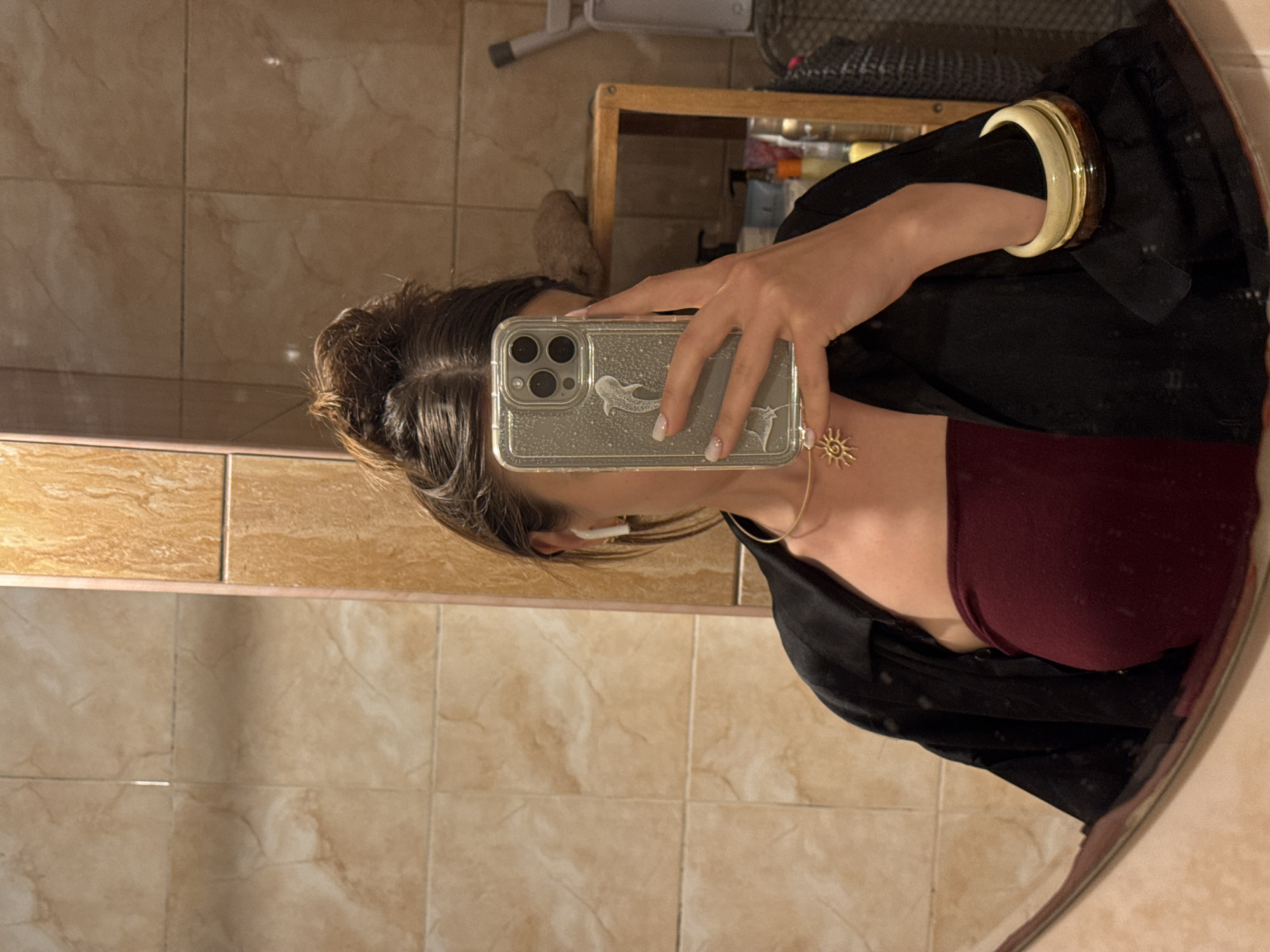 A person is taking a mirror selfie in a bathroom. The person has their hair in a bun and is wearing a black open shirt over a maroon strapless top. They are also wearing a gold sun necklace and bangles on their wrist. The bathroom has tiled walls.