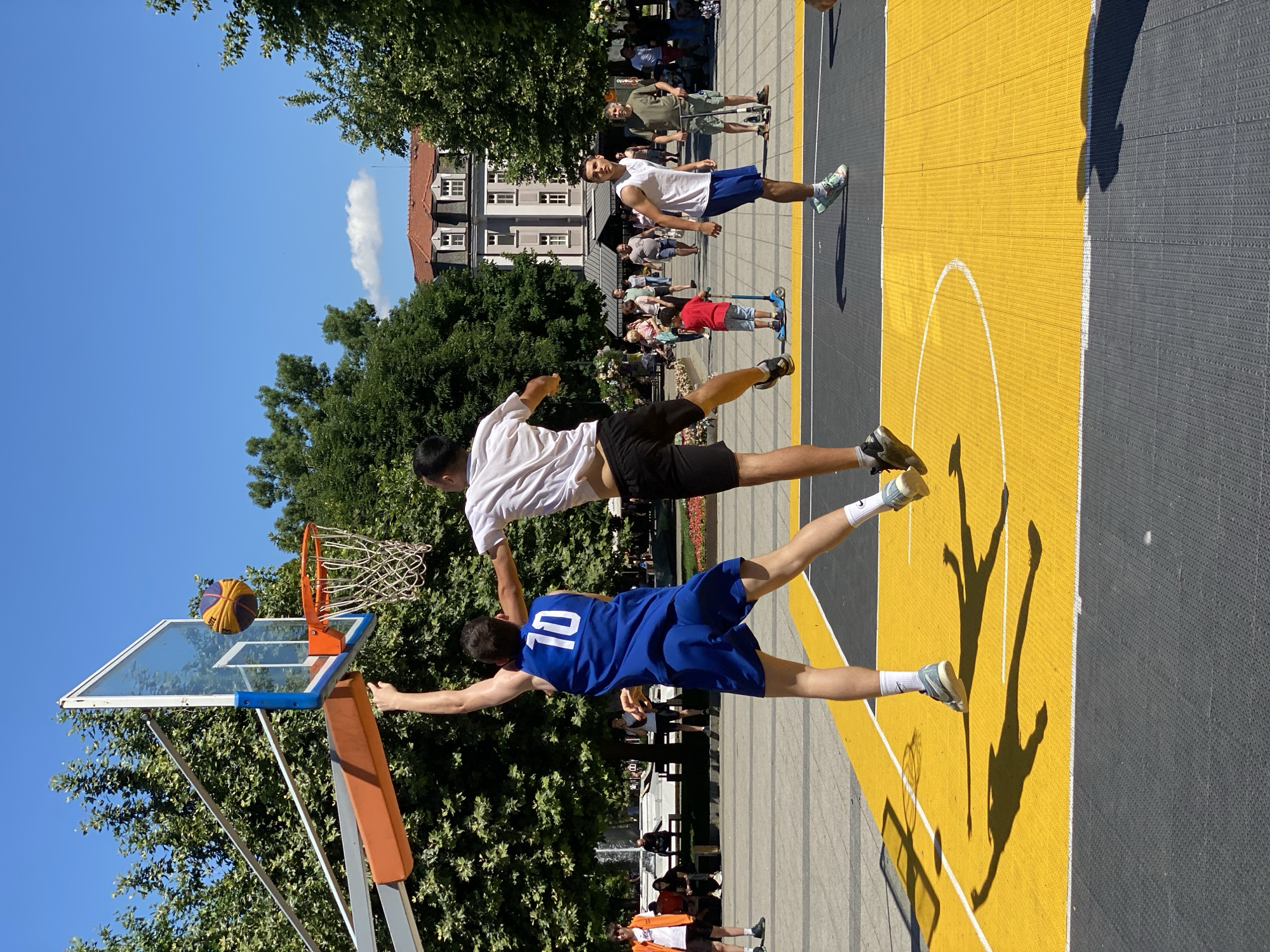 The image depicts a sunny day scene on a basketball court in Pleven, Bulgaria, likely in the area of the City Garden. Two young men are actively playing basketball. One player, wearing a blue jersey with the number 10 and blue shorts, is in mid-air, reaching for the basketball, which is nearing the hoop. He is being defended by another player in a white t-shirt and black shorts, who is also jumping and attempting to block the shot. The basketball court itself is marked with yellow and black surfaces. In the background, there are trees, people walking, buildings with red roofs, and a fountain. The overall atmosphere suggests a casual, outdoor sporting activity in a public urban space.