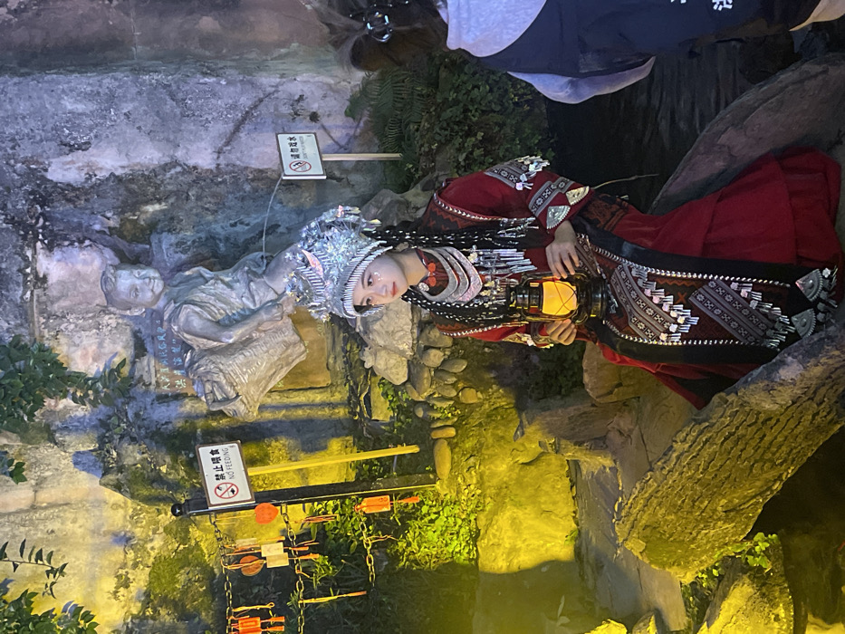 An outdoor scene in Yuzhong District, China, depicts a young female in elaborate traditional attire seated on a log, holding a lit lantern. She wears a red dress with intricate patterns and silver-colored embellishments, a multi-layered silver necklace, and a large, ornate silver headdress. Glitter is applied to her upper cheeks. Her gaze is directed left.
Behind her, against a rocky backdrop, a statue of a young boy carrying a basket is visible, serving as a fountain with water appearing to emanate from its lower torso. To the statue's left, a "NO FEEDING" sign in Chinese and English is posted on a black pole. To the statue's right, a "DON'T PLAY WATER" sign, also in Chinese and English, is visible. The rocky background features green foliage and is unevenly lit; the left side has a yellow-green illumination, while the right side is darker with cooler tones. Several red and rectangular wish-like charms hang on chains to the left of the "NO FEEDING" sign. Dark water is visible at the base of the rocks and in the lower right foreground. Partially visible on the far right is another individual, facing away, wearing a grey hoodie over a dark garment.