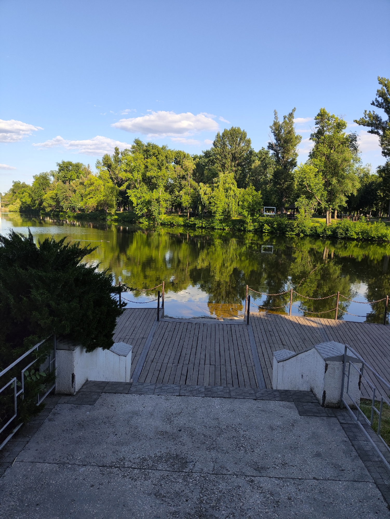 The image shows a calm body of water, likely a river or lake, with reflections of lush green trees lining its banks. A wooden pier extends into the water from the foreground, which features concrete steps leading down to it. The sky is a clear blue with a few scattered white clouds. The scene appears to be a park or recreational area, possibly in the city of Kryvyi Rih, Ukraine, given its typical landscape features. There are no people or animals visible in the image. The overall atmosphere is peaceful and serene.