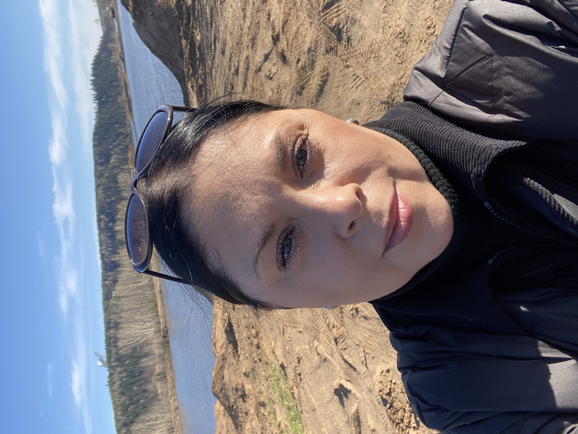 A woman with dark hair and fair skin takes a selfie outdoors on a sunny day. Sunglasses rest on her head. She wears a black turtleneck sweater and a black puffy jacket. The background shows a sandy area with vehicle tracks, a body of water, and trees. The location is likely Noviy, Russia.