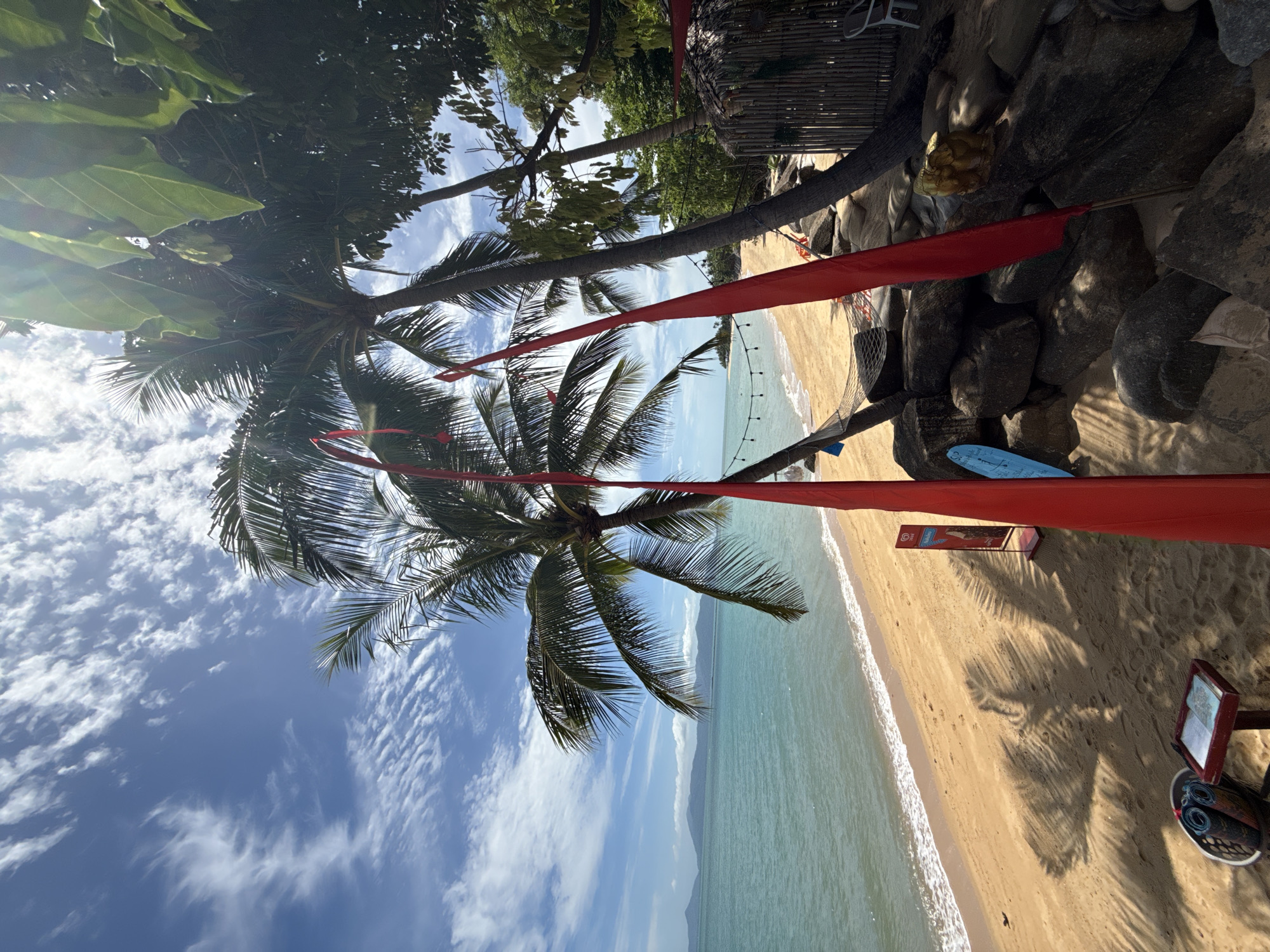 The image depicts a tropical beach scene in Baan Bo Phut, Thailand. The foreground features sandy ground with shadows of palm trees cast across it. A small red table with a basket containing rolled mats and a menu is visible on the left. Two bright red fabric banners hang vertically, obscuring parts of the scene. In the middle ground, a net hammock is suspended between palm trees, and a cluster of large rocks sits behind it. To the right, a rustic hut with a thatched roof and bamboo walls is partially visible, with a white chair outside. A golden statue of what appears to be an elephant-headed figure rests on the rocks. The background shows a tranquil turquoise ocean meeting a sandy beach, with a line of palm trees and vegetation along the shore. In the distance, several islands or peninsulas are silhouetted against a blue sky filled with wispy white clouds. The sun is bright, creating lens flare in the upper right portion of the image.