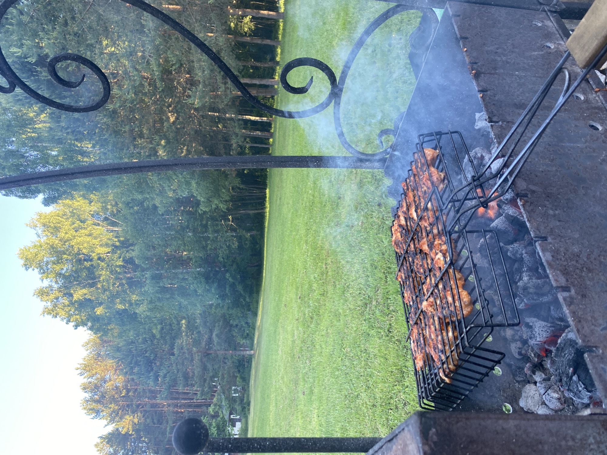 The image depicts an outdoor grilling scene in the city of Копнино, Russia. In the foreground, a metal brazier contains glowing charcoal embers and ash. A black metal grilling basket, holding multiple pieces of meat (appearing cooked with some charring), is positioned directly over the coals. Smoke emanates from the grill, partially obscuring the scene. The brazier features decorative metal scrollwork on its right side and a vertical metal post on the left. A wooden-handled implement, likely part of the grilling basket, extends from the lower right. Beyond the grill, a large, expansive green lawn occupies the midground. In the background, a dense forest line consists of a mix of coniferous and deciduous trees, with sunlight illuminating their upper foliage. A small, white building with a dark roof is partially visible among the trees on the left side of the distant lawn. The sky above is clear and light blue. No individuals are present in the frame.