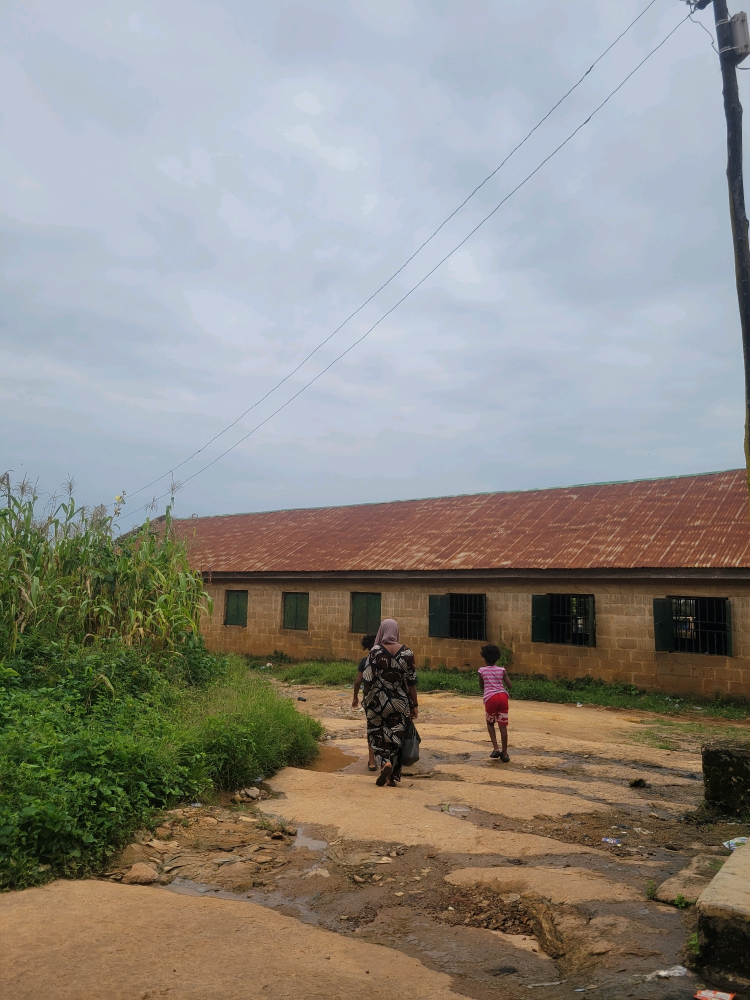 In Jos, Nigeria, a group of three individuals, appearing to be two adults and one child, walk away from the camera down a dirt road. The adult on the left wears a patterned dress and carries a bag. The child, on the right, wears a striped pink shirt and red patterned shorts. They are walking in front of a long, single-story building constructed of pale bricks with a rusty, corrugated metal roof. The building has several rectangular windows, each covered with dark green shutters and bars. To the left of the building, a dense patch of tall green corn stalks grows. The road itself is uneven, with puddles of water and debris scattered on its surface, suggesting recent rain or poor drainage. Overhead, a utility pole stands to the right, with wires stretching across the cloudy sky. The overall scene depicts a rural or semi-urban environment in Jos, characterized by simple architecture and natural elements.