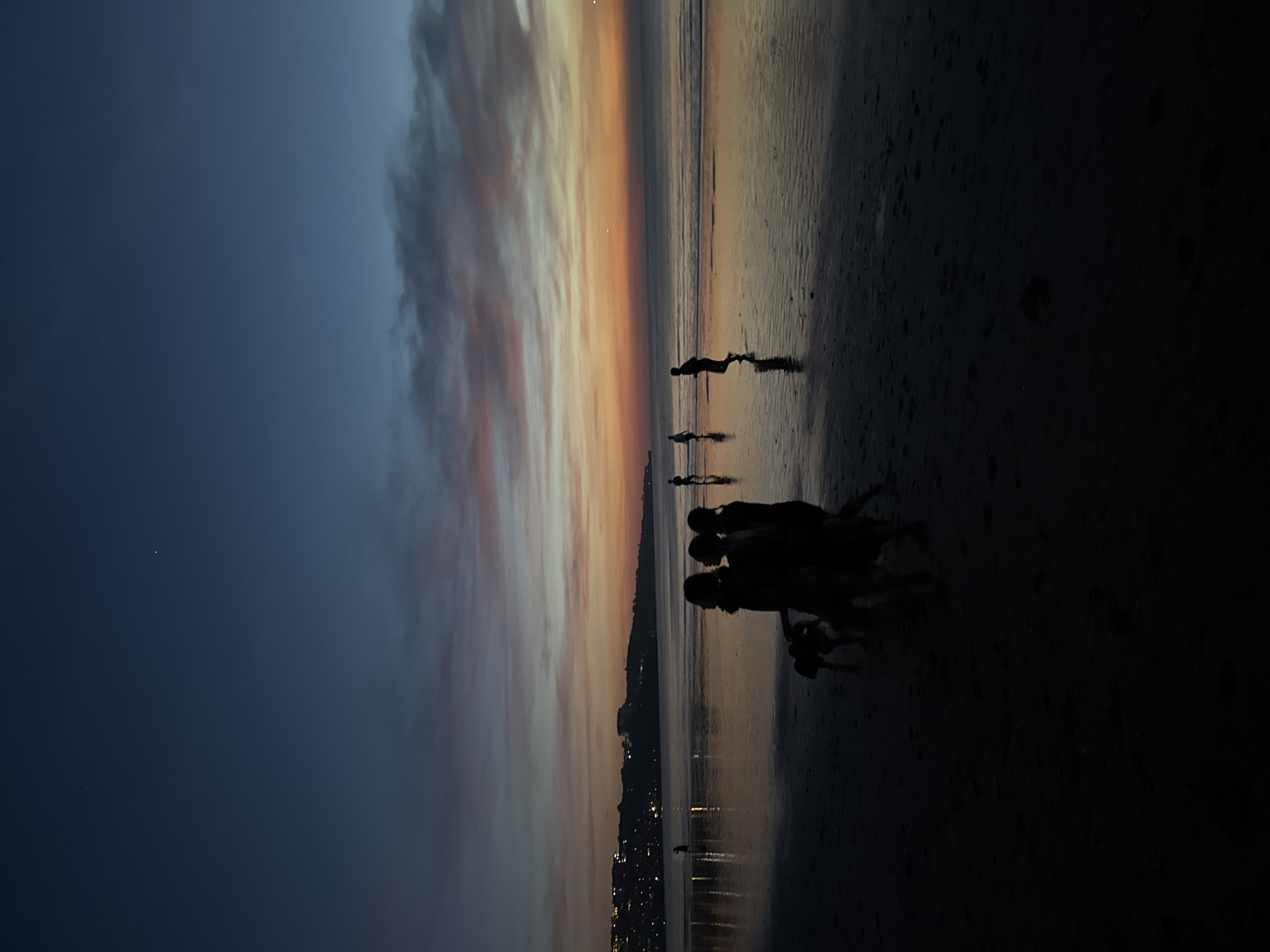 Captured in Kuta Selatan, Indonesia, the scene unfolds on a beach at dusk, with the skyline silhouetted against a sunset that transitions from dark blue to orange. The city lights twinkle along the shoreline. Several people, also silhouetted, are gathered near the water's edge, while another individual is further out, seemingly walking or running along the wet sand. The wet sand reflects the colors of the sky and city lights.