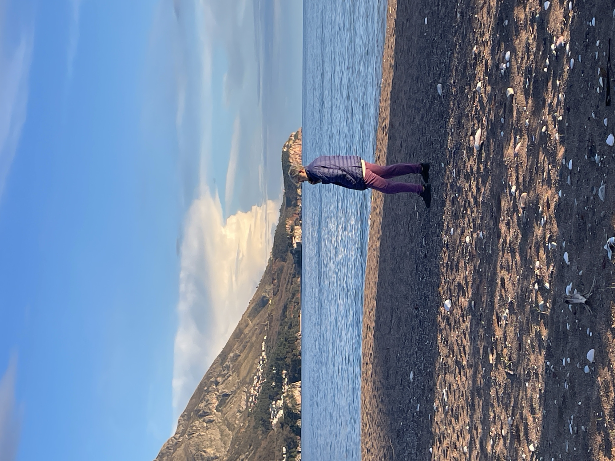 The image displays a coastal scene in Bečići, Montenegro, under a partly cloudy sky, likely during the late afternoon. The foreground features a dark pebble and sand beach, sparsely covered with scattered light-colored stones and small pieces of dark organic debris.
A single individual, an adult, stands on the beach, positioned slightly left of center. The person is observed from a rear-quarter angle, facing towards the right side of the frame, gazing towards the distant sea and coastline. They are dressed in a dark blue puffer jacket, maroon or purple trousers, and dark shoes, with a light-colored collar visible beneath the jacket.
Beyond the beach, the calm, blue-green waters of the sea extend across the midground to the horizon. The background is dominated by a rugged, verdant mountain range rising steeply from the coast on the left. Numerous white structures, consistent with a coastal settlement, are visible clustered on the lower slopes and base of this mountain. A distinct rocky headland projects into the sea on the right side of the distant view. The sky above is clear blue with scattered white and grey clouds; a prominent, bright white cumulus cloud formation is visible above the right headland. Shadows on the beach indicate a low sun angle from the left.