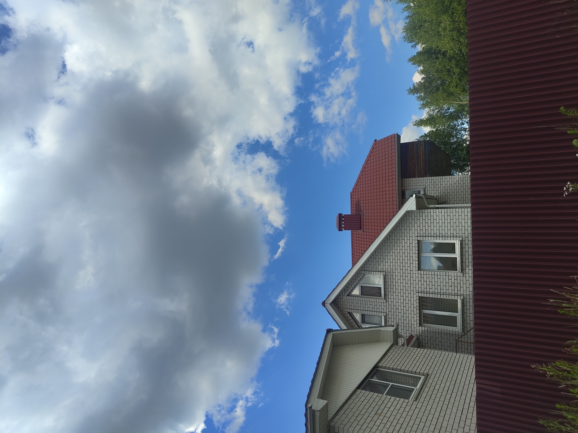 The image shows a view of residential buildings and a fence under a cloudy sky. In the foreground, a rust-colored corrugated metal fence runs horizontally across the frame. Behind the fence, two residential buildings are visible. The building on the left has a white, angled roof and is constructed with light-colored bricks. The building on the right features a red, pitched roof with a brick chimney and is also made of light-colored bricks, with several windows visible. A line of green trees is visible behind the right building, and the sky above is a mix of blue sky and large, grey clouds. The location is identified as Tsibino, Russia.
