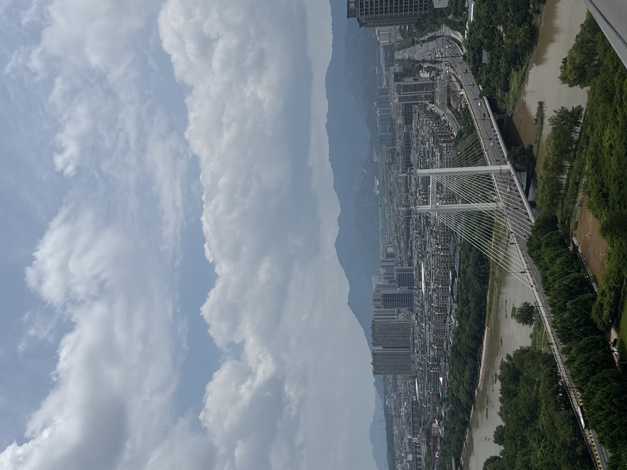 The image depicts a wide-angle view of a city, likely Yiwu, China, on a cloudy day. In the foreground, a modern cable-stayed bridge spans a brown river, with lush green trees lining the riverbanks. The bridge has multiple lanes of traffic, with cars and a bus visible. Beyond the river and bridge, a dense urban landscape unfolds, characterized by numerous apartment buildings and other structures. In the distance, a range of mountains forms the backdrop to the city. The sky is filled with white and gray clouds, suggesting overcast conditions. There are no discernible individuals in focus, and the scene is dominated by urban infrastructure and natural elements.