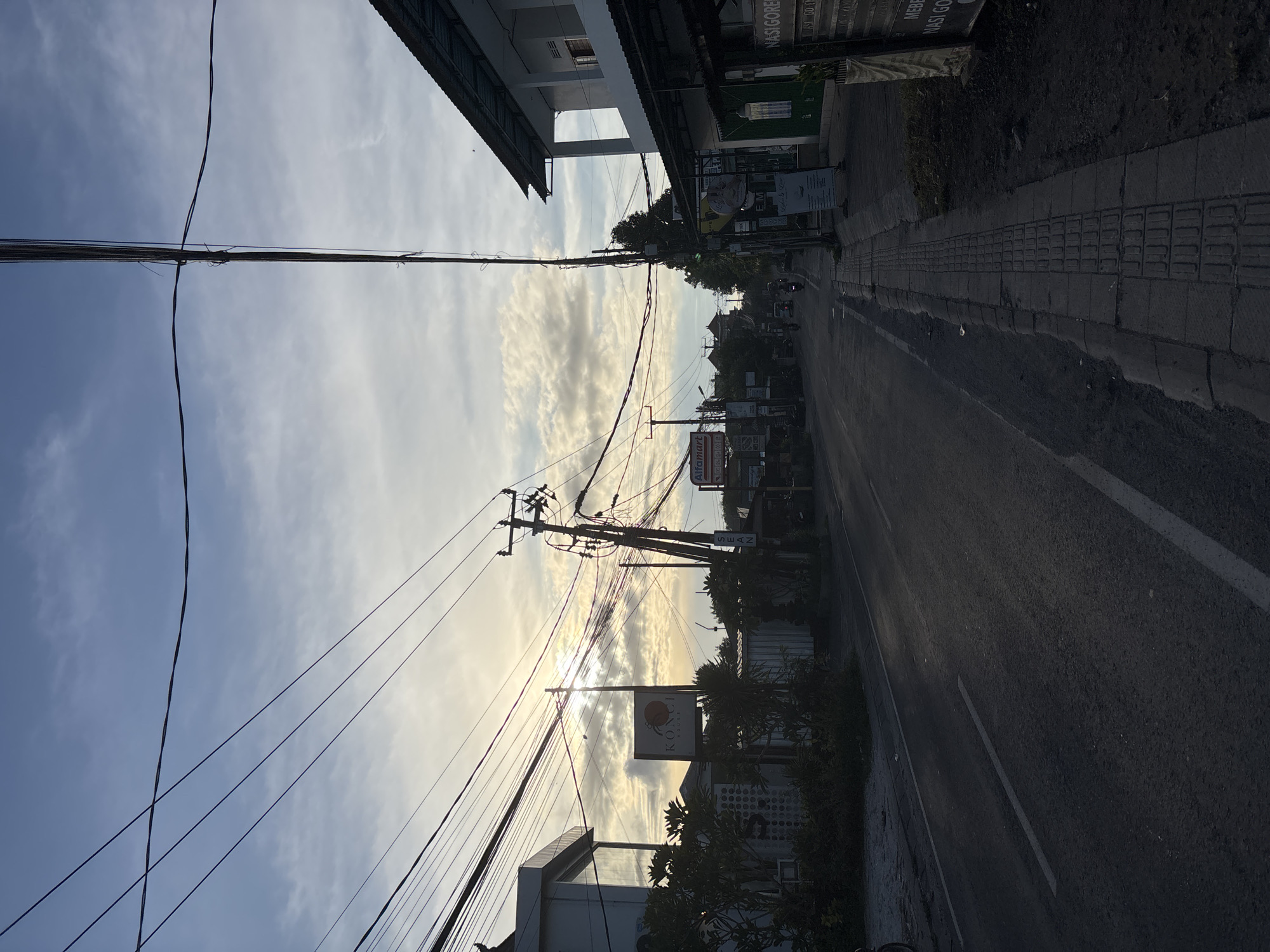 The image depicts a street scene in Ungasan, Indonesia. The view is along a narrow road with buildings lining both sides. Overhead, a dense network of power lines crisscrosses the sky, partially obscuring a sky filled with clouds. Along the right side of the road, a paved sidewalk with tactile paving is visible. The road surface appears asphalt, with white lane markings. A sign for "KONCI House" is seen along the lower-right side of the street. The sun appears to be setting or rising, casting a glow through the clouds. There are cars and motorcycles on the street.