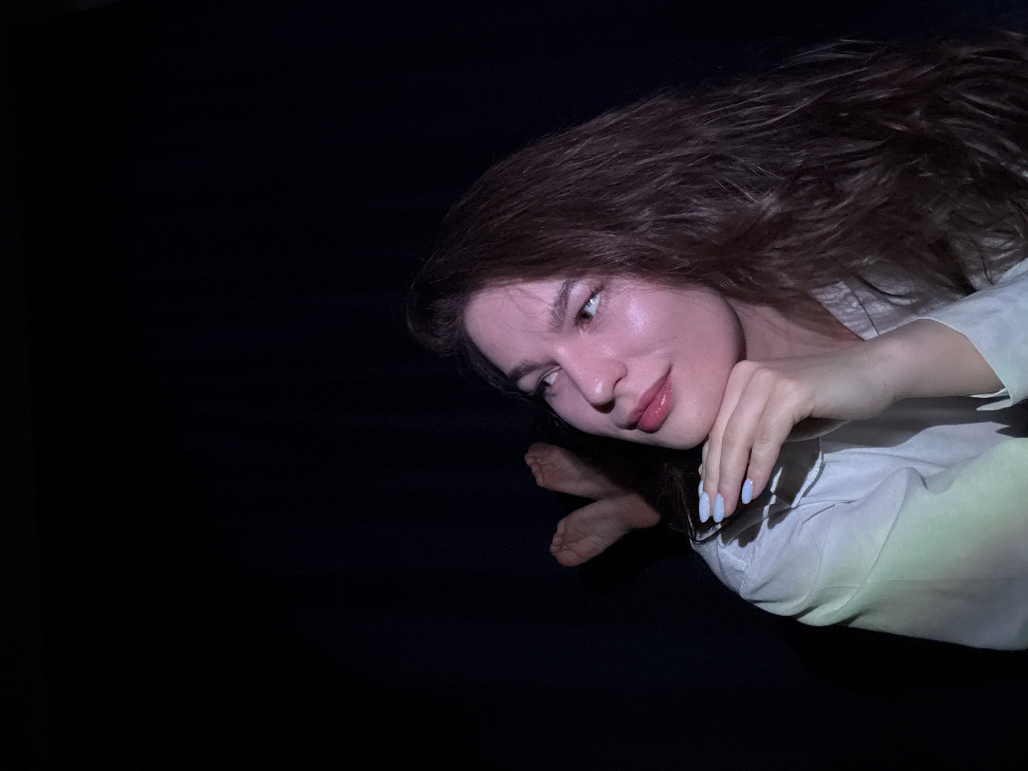 A young woman with long brown hair and fair skin is positioned in the upper right quadrant of the image, set against a dark background. She is wearing a white, collared shirt. Her face is illuminated, revealing smooth skin and a gentle expression as she gazes toward the left. Her left hand is gently resting near her face, displaying pale blue nail polish. Below her face, two bare feet are visible. The environment appears to be indoors due to the controlled lighting. There is no information available to identify the location as Chaikovsky, Russia, based solely on the image.