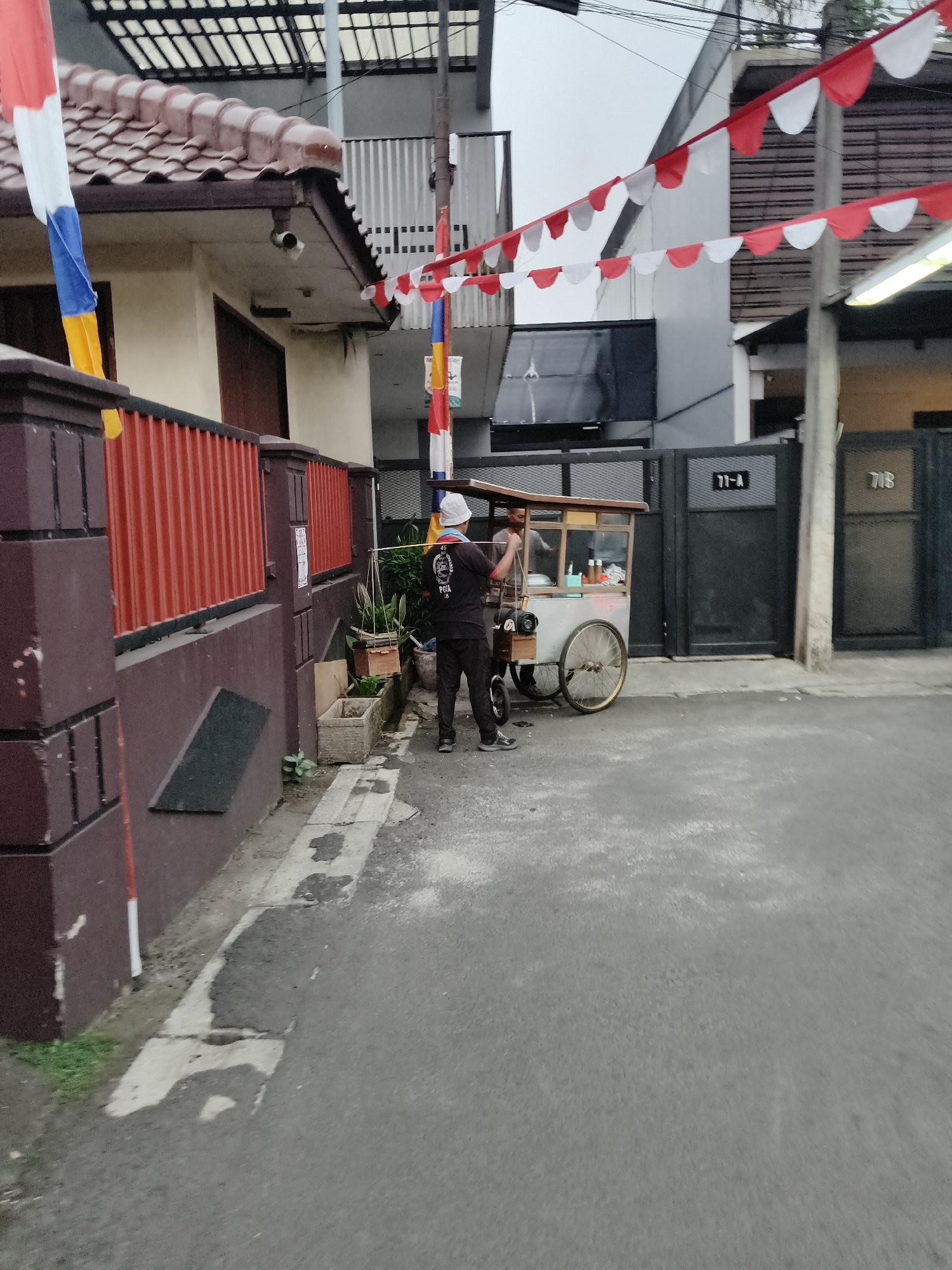 The image depicts a street scene in South Tangerang, Indonesia, possibly during a festive or commemorative occasion, evidenced by the red and white triangular flags strung across the street and on a pole. In the foreground, a two-person food cart with a wooden top and metal frame is parked on the asphalt road. One person, wearing a white hat and a dark t-shirt with white text, stands behind the cart, facing away from the camera, appearing to be serving a customer. The second person, partially obscured, is likely the customer. A red and white striped pole is visible to the right of the food cart. To the left, a building with a tiled roof and a red corrugated fence is present. Further back, another building with a grey facade and large dark gates, marked with numbers "71-A" and "71B", is visible. The overall environment appears to be an urban street with residential or commercial buildings.