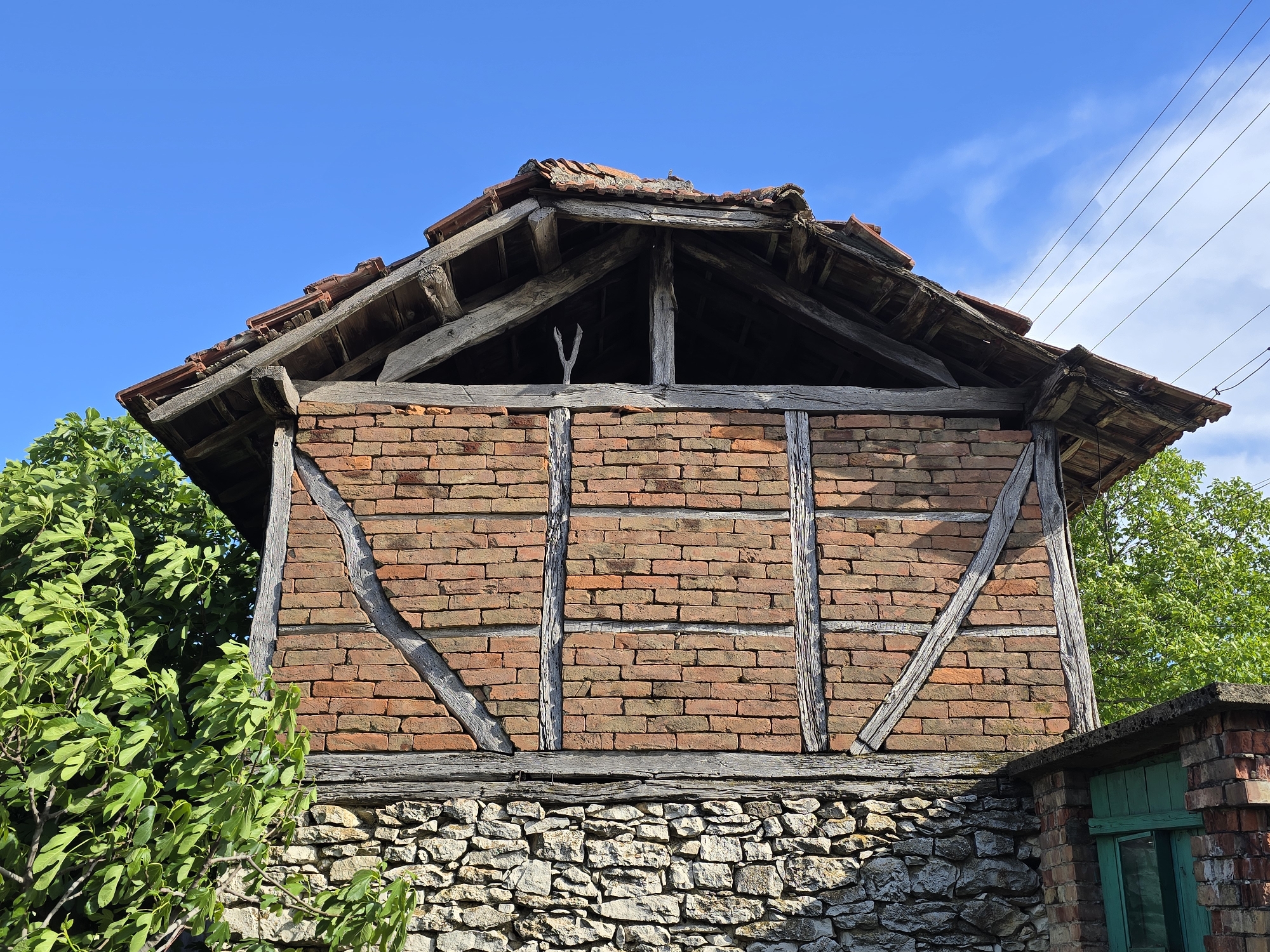 A low-angle, medium shot depicts the gable end of a small, decaying timber-frame structure built on a stone foundation under a clear blue sky. The location is noted as Unknown, Unknown.
The lower portion of the building is constructed of roughly hewn stones mortared together, forming a retaining wall-like base. Above this, the timber frame is filled with red bricks laid in a simple pattern. Gray, weathered timbers form the structural support, including diagonal braces.
The roof is gabled, with a worn and uneven red tile covering. The roof's timber frame is exposed, showing signs of age and decay. A forked branch is placed in the gable apex.
The structure is surrounded by foliage. To the left, a tree with green leaves partially obscures the lower left corner. A similar tree is on the right side. A small, brick structure with a green-painted window and door is attached to the right side of the main building. Electrical wires run diagonally across the upper right portion of the sky.
